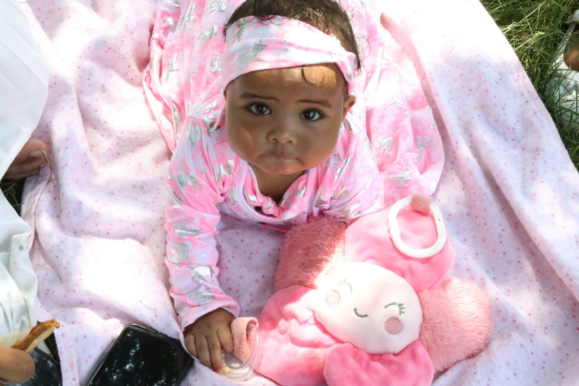 Baby in pink outfit and headband, looking forward on a pink blanket, holding a pink plush flower.