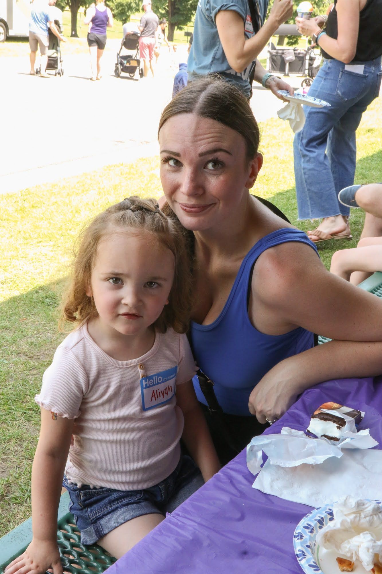 Woman and child seated at a picnic table. Woman in blue tank top; child in pink shirt and denim shorts. Smiling.