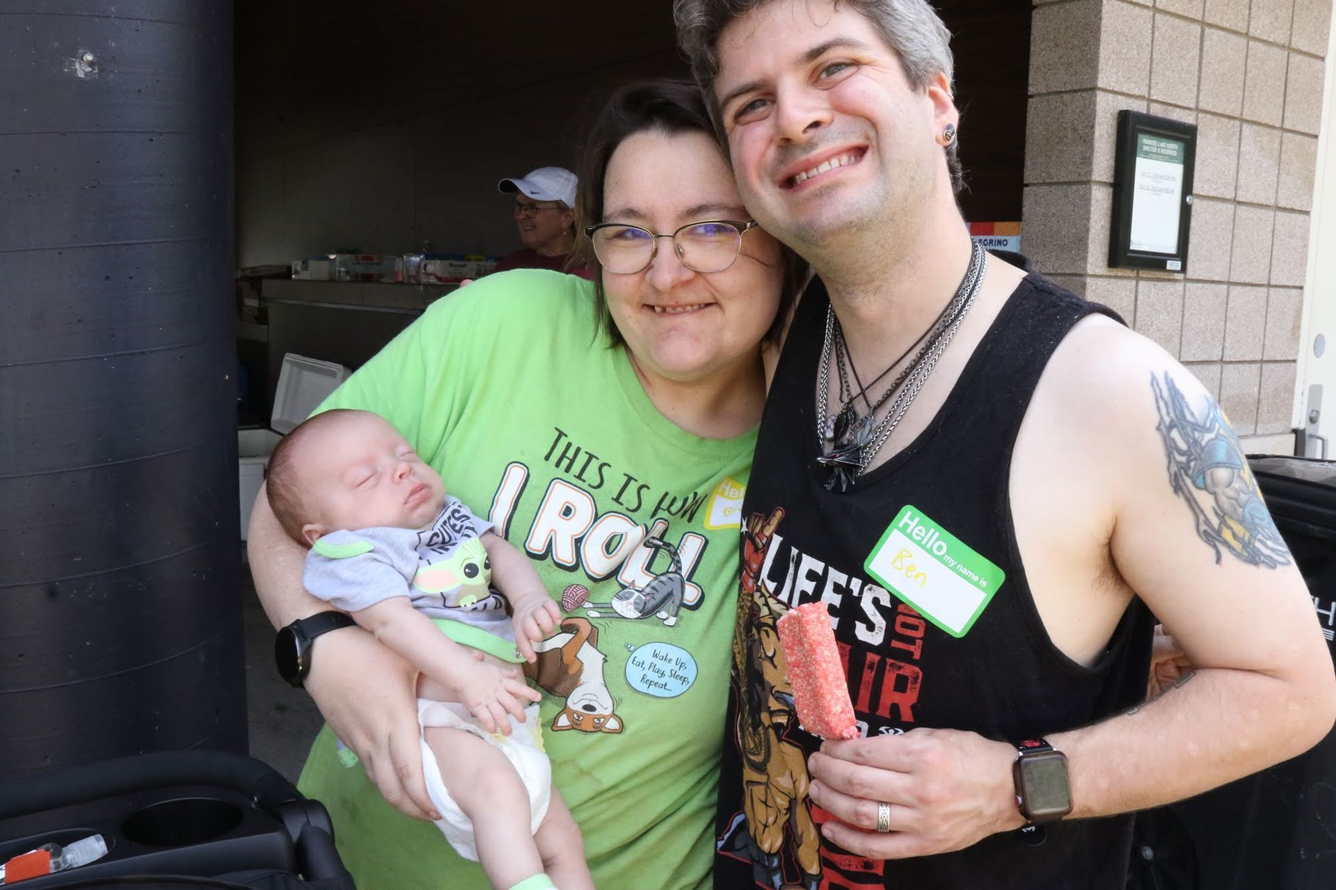 Couple and baby smiling outside. Woman in green shirt holds the baby. Man holds a popsicle.