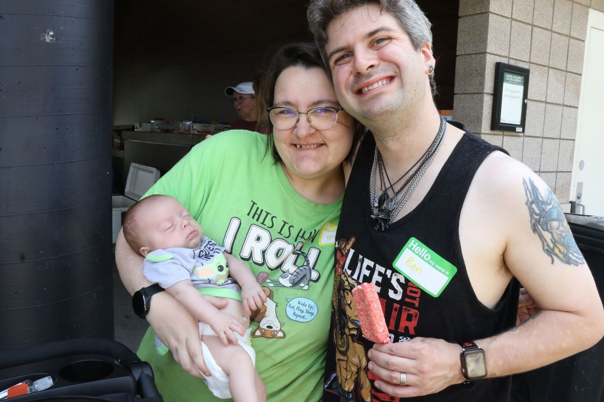 Woman holding a sleeping baby smiles next to a smiling man with a tattoo, both outside.