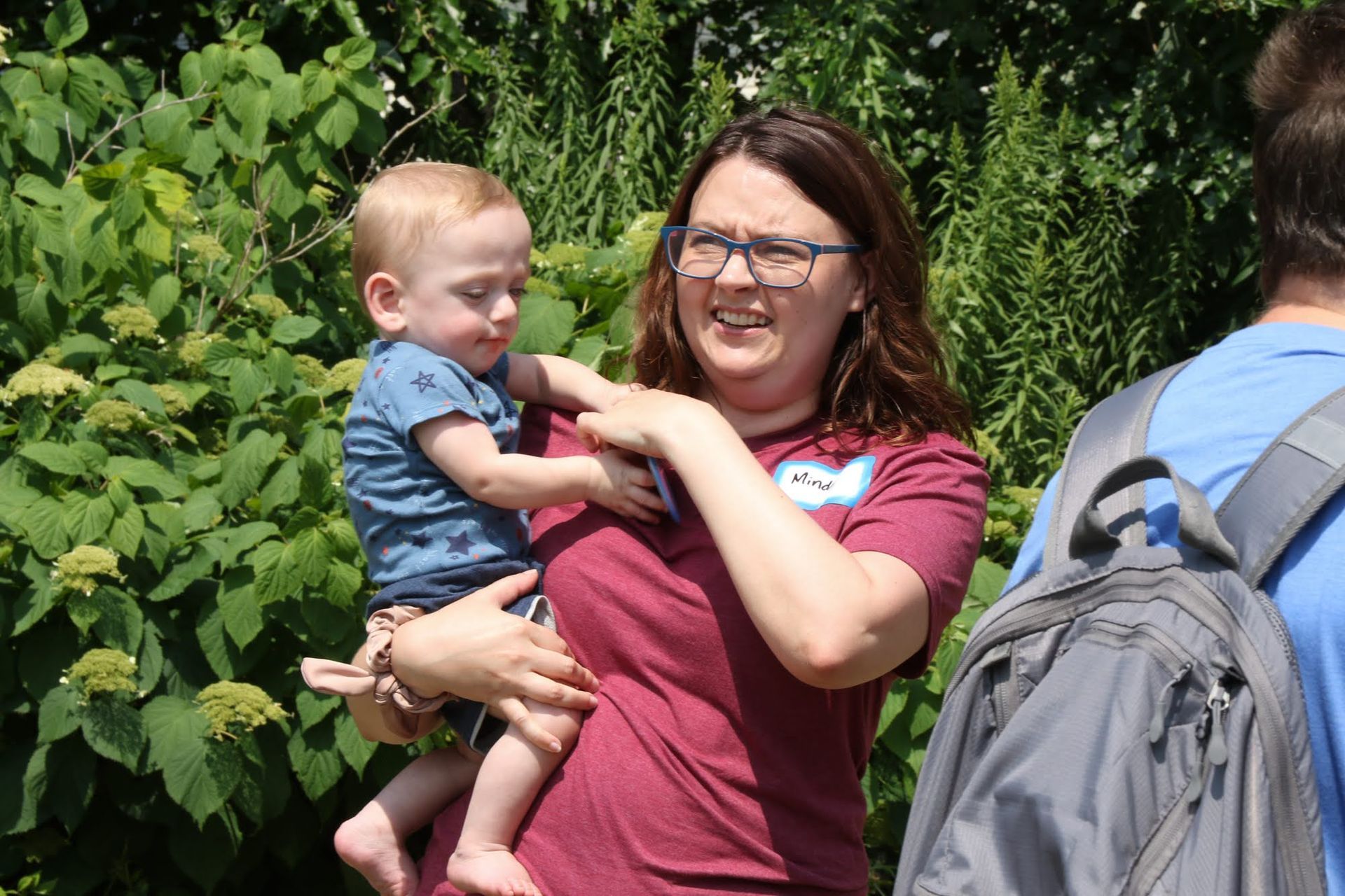 Woman in maroon shirt holds a toddler in front of green bushes; a person with a backpack stands nearby.
