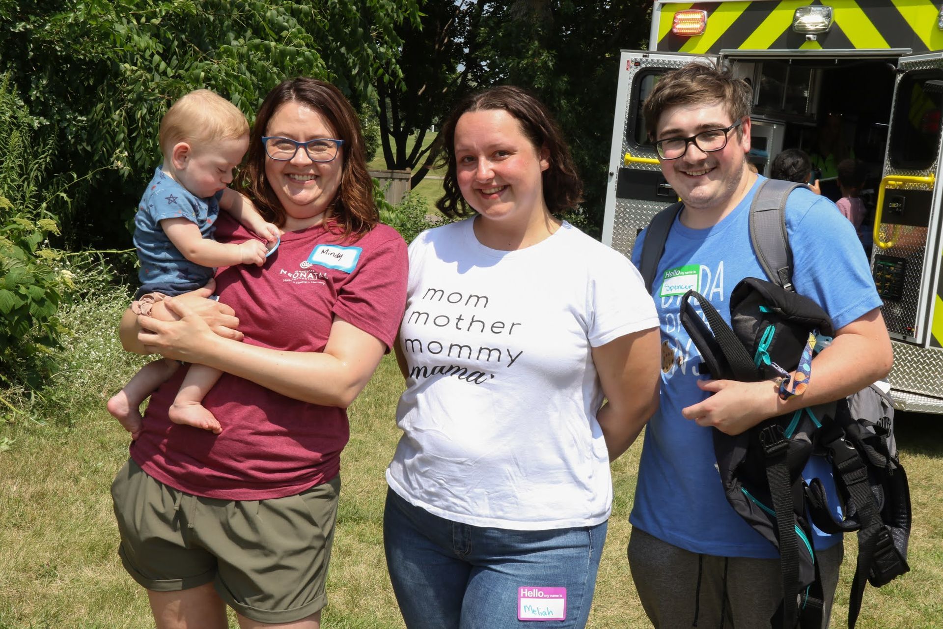 Three adults and a baby pose outdoors near an ambulance. One adult holds the baby; another wears a 