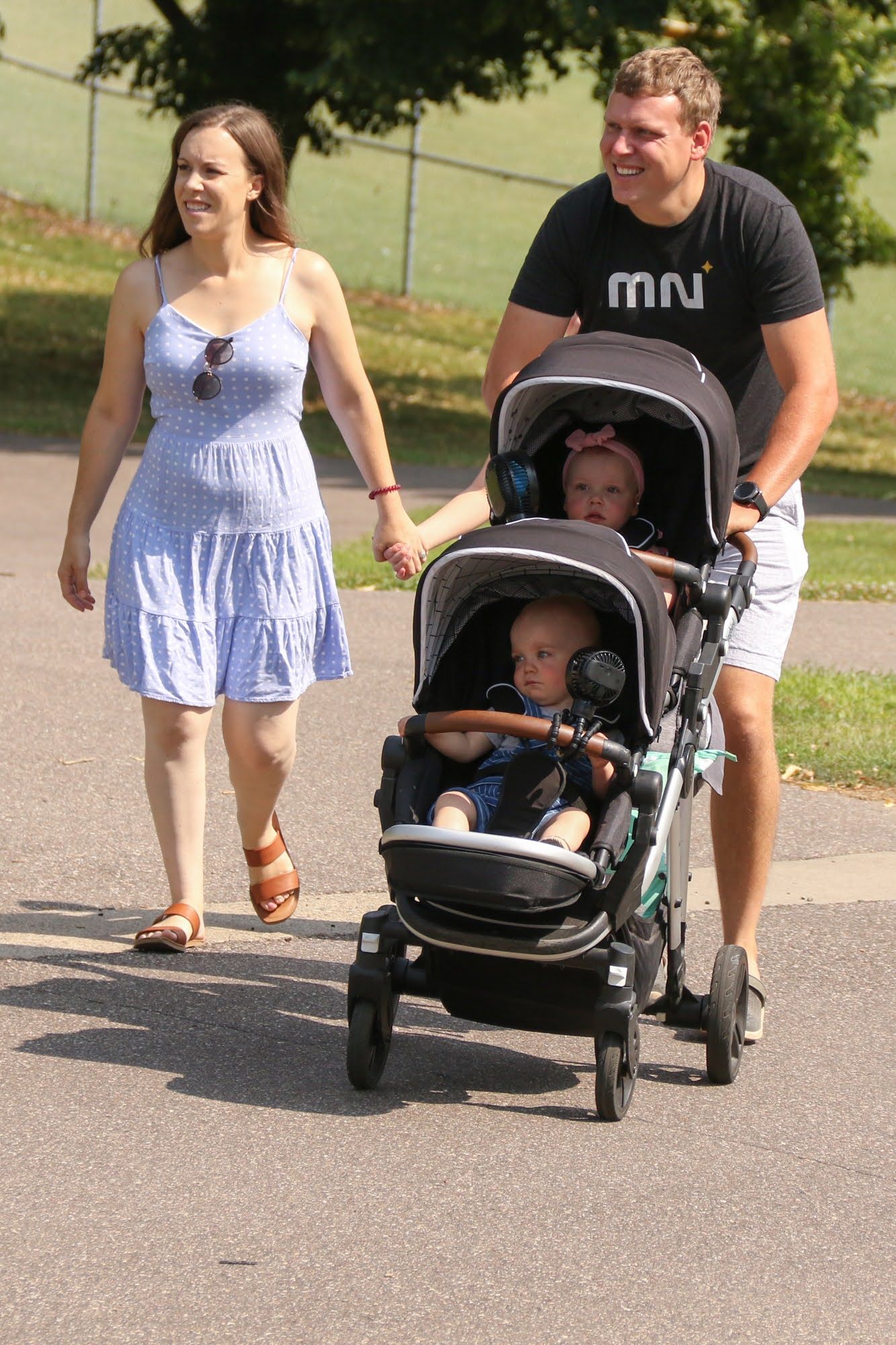 Parents push a double stroller with two babies. Woman holds hands and smiles, man smiles. Outdoors.