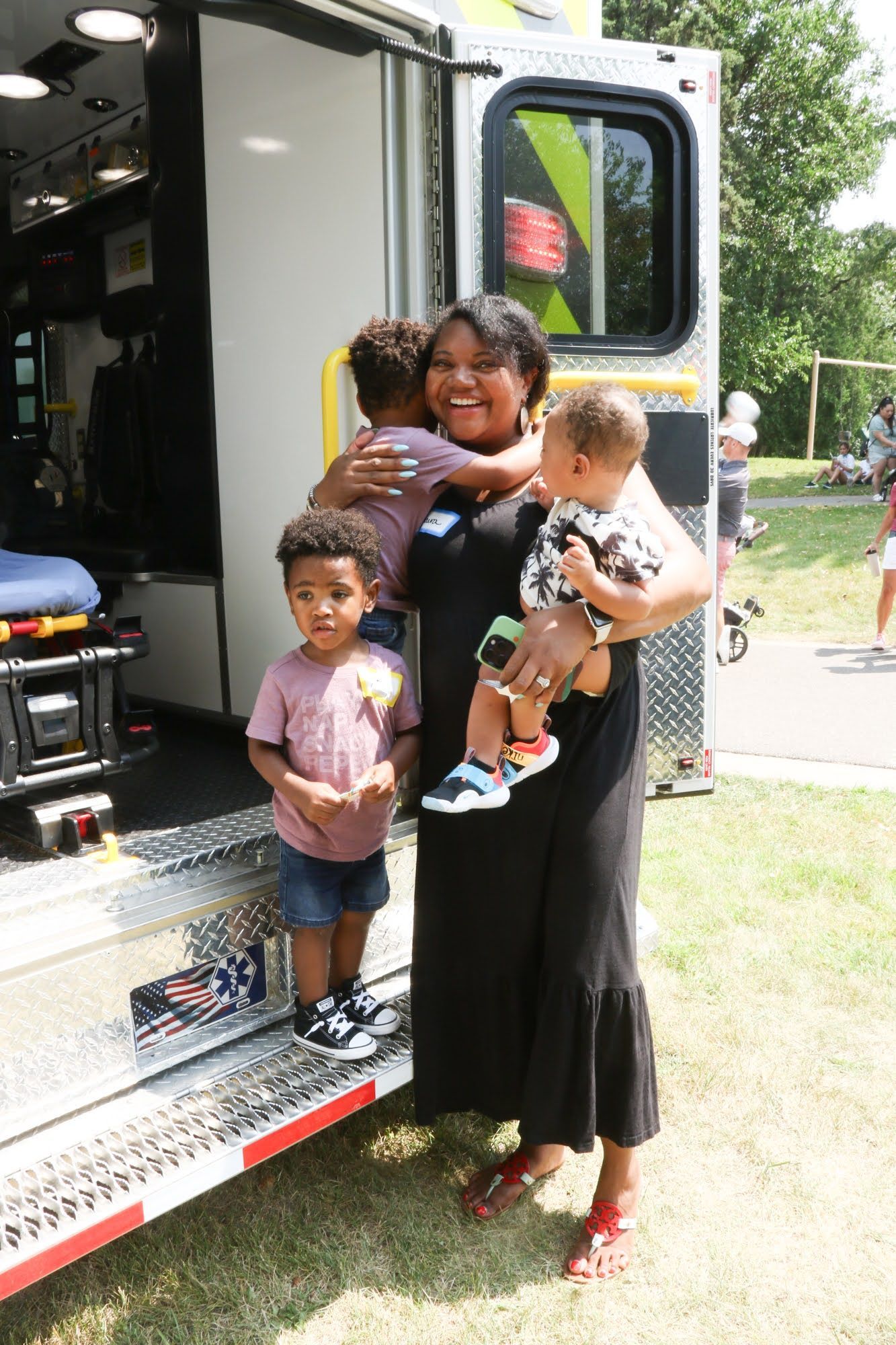 Woman holding two children in front of an ambulance, smiling. Outdoor setting, sunny day.