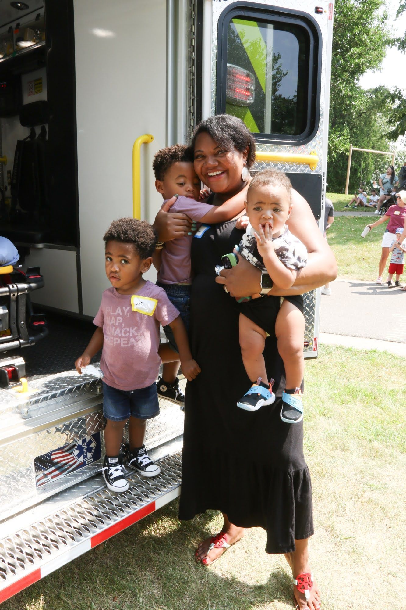 Woman smiles, holding two young children while another stands nearby an ambulance. Outdoor setting.