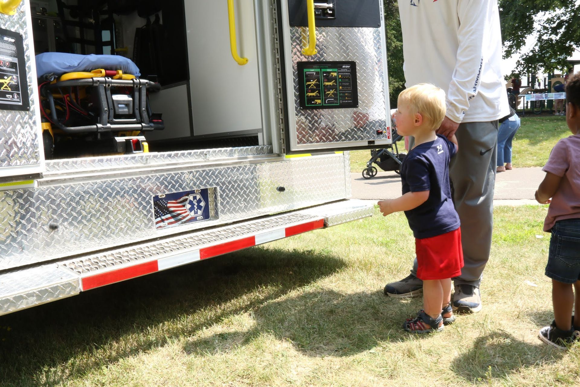 Child in red shorts looks at an ambulance with an adult nearby. Other children are visible. Outdoors.