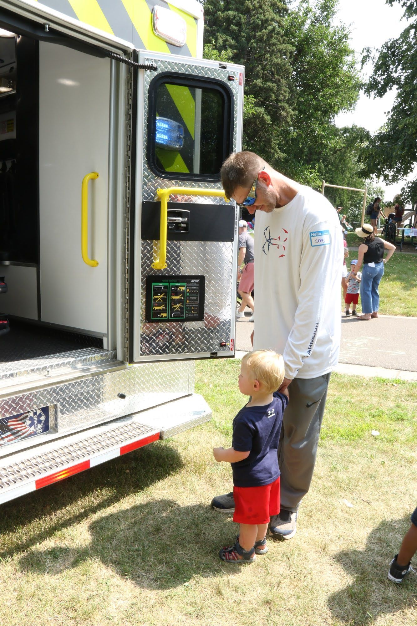 Man and child looking at the open back of an emergency vehicle; grass and trees in background.