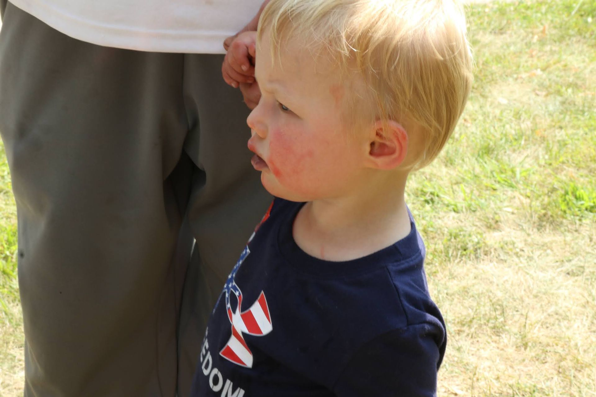 Blond toddler with stained face looks to the side while standing near someone in khaki pants and a lawn in the background.