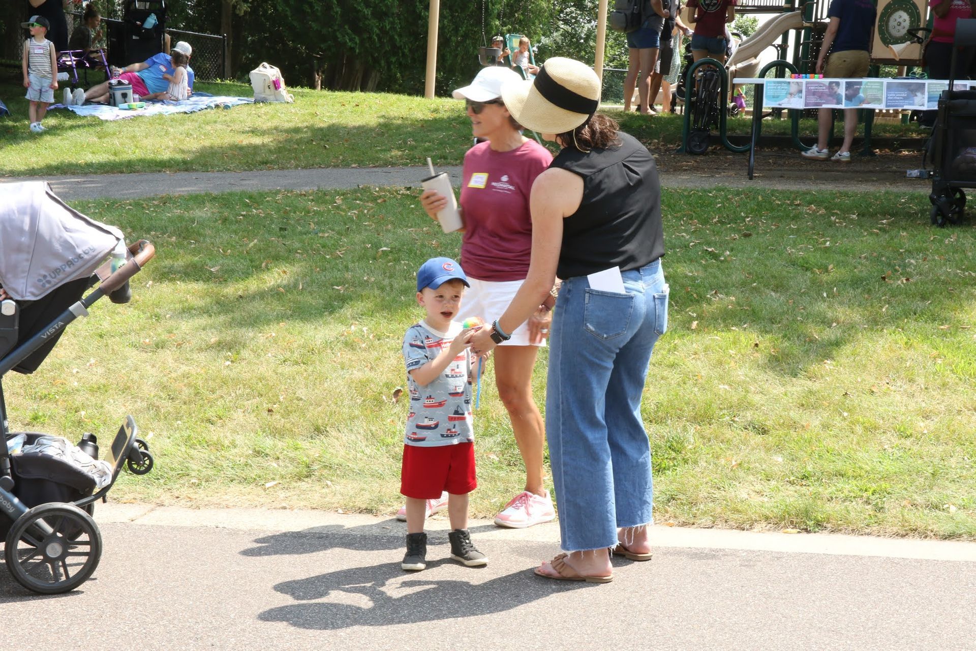 Boy in red shorts and blue cap with two women on a path in a park; stroller on left.