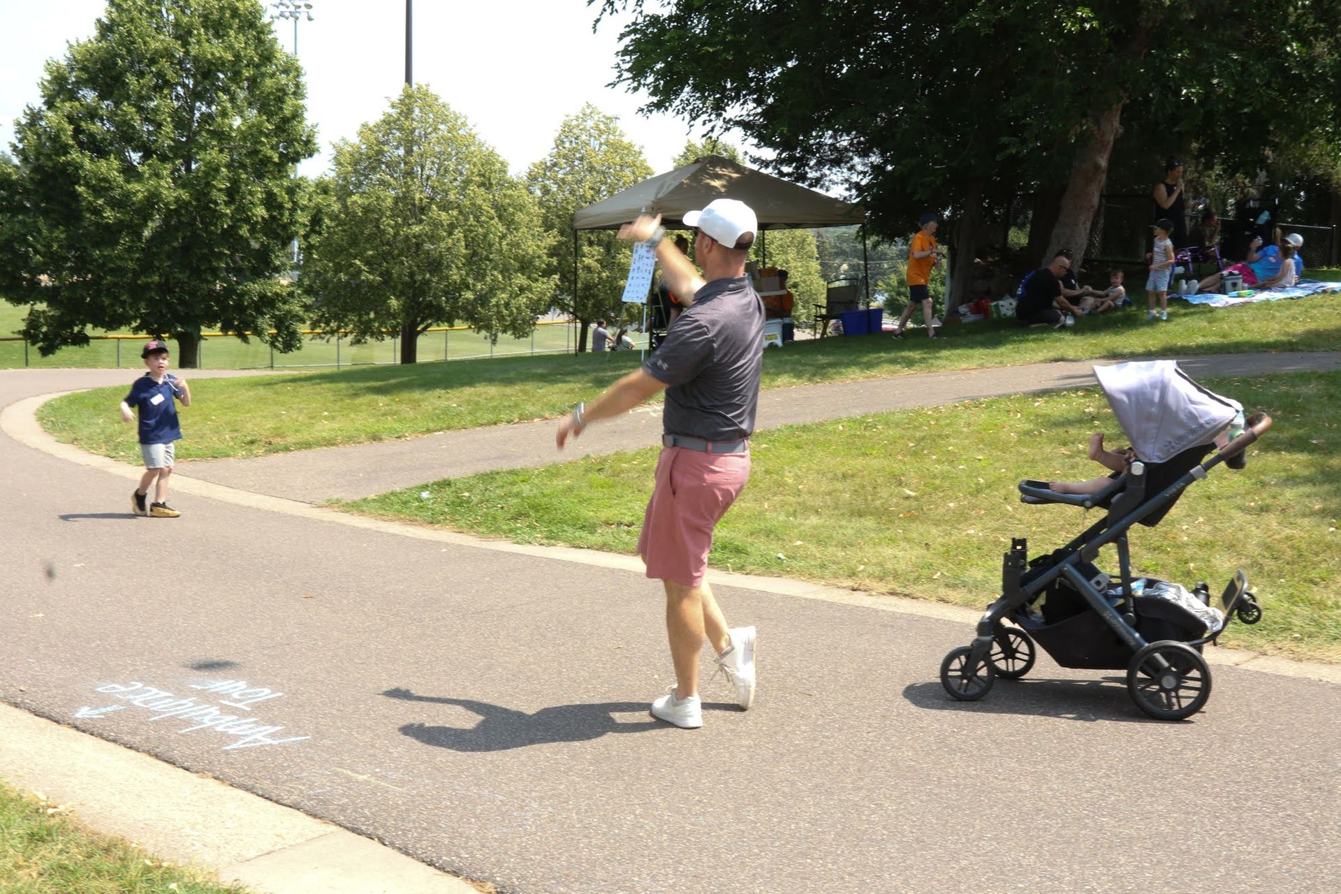Man throwing water, son watching, stroller nearby, park setting, sunny day.