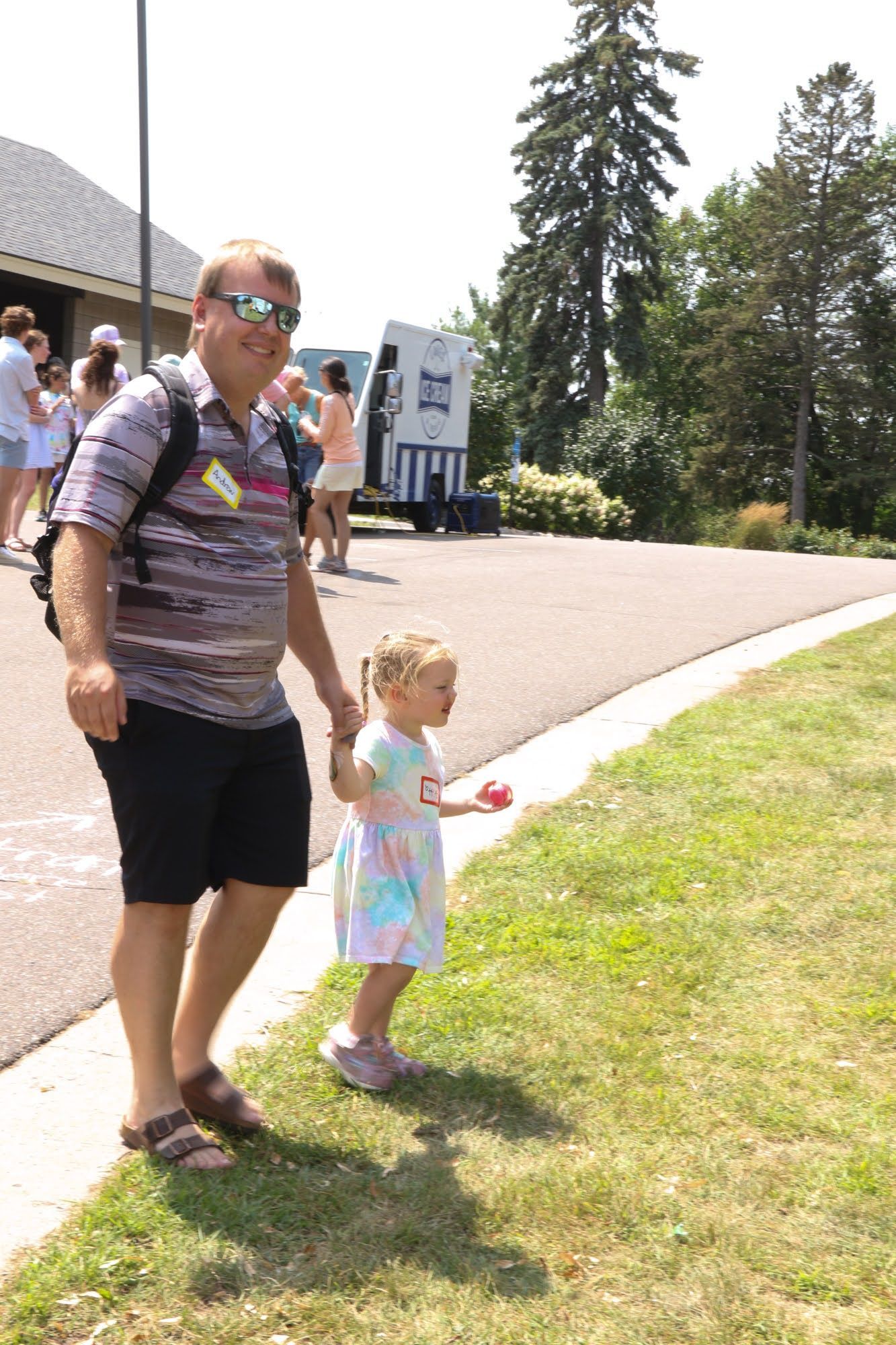 Man and small child walk on grass path. Man holds child’s hand near an ice cream stand.