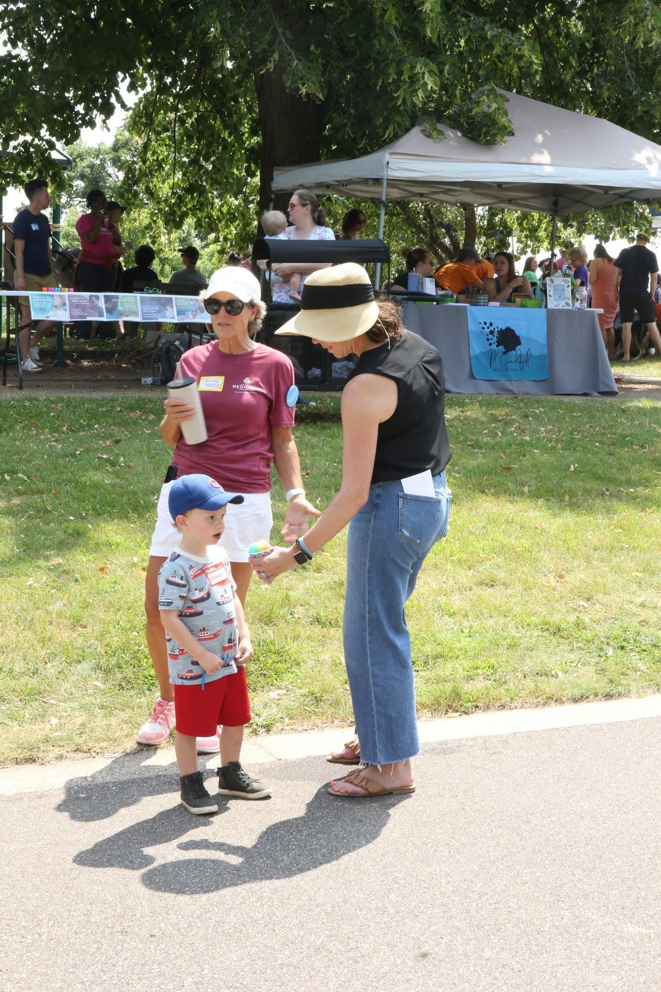 Woman in a hat and jeans interacts with a child wearing a hat and red shorts at an outdoor event.
