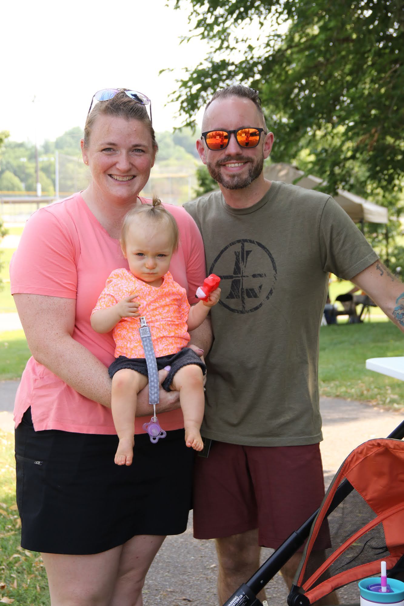 Woman holding a baby, next to a man; all are smiling outdoors near a stroller.