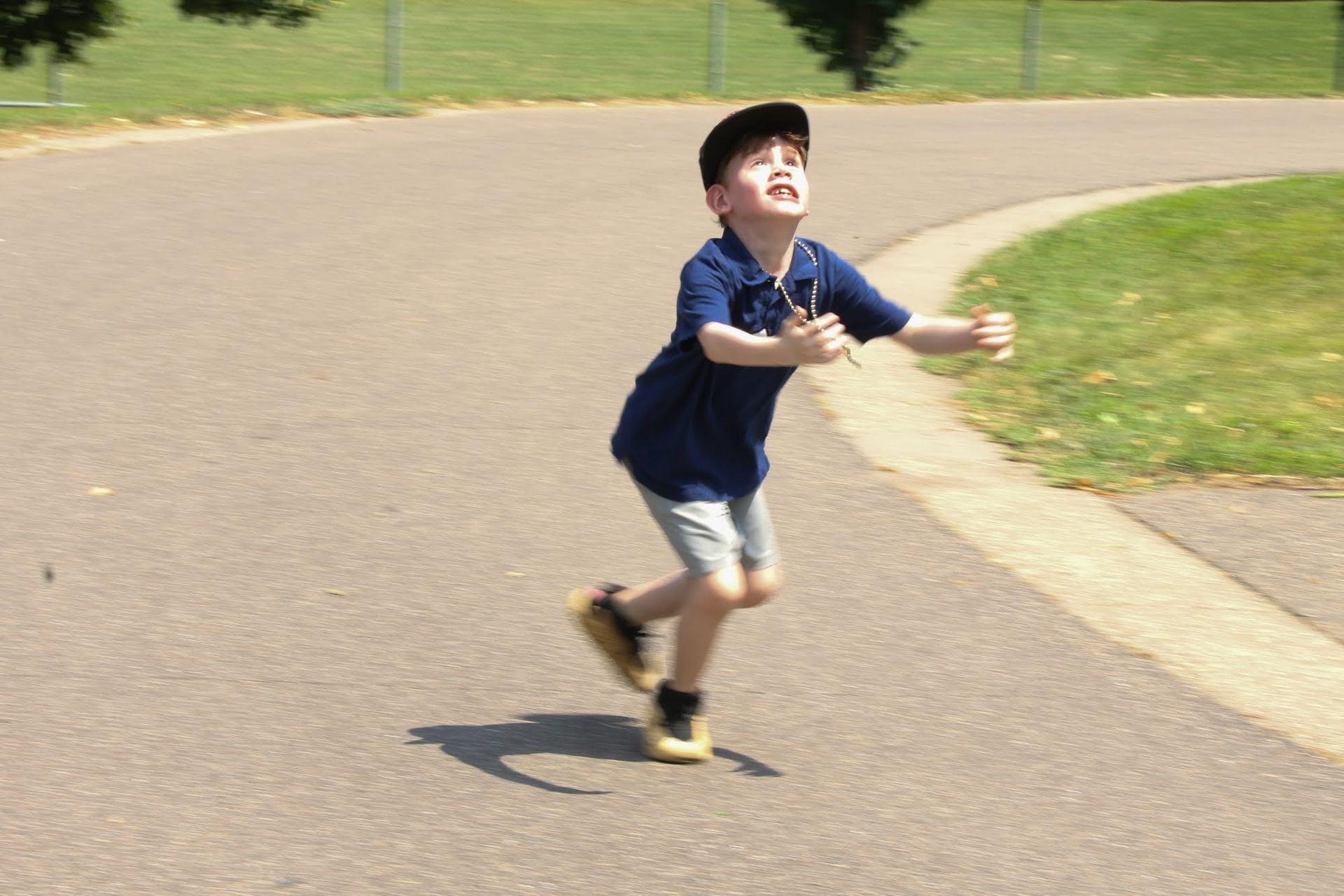Boy in a navy shirt and cap runs on a paved path, arms outstretched, looking upwards.