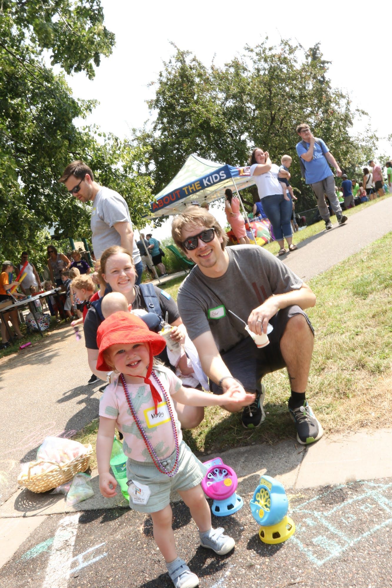 Child smiles, wearing red hat, plays with bubbles, with adults nearby in park setting.