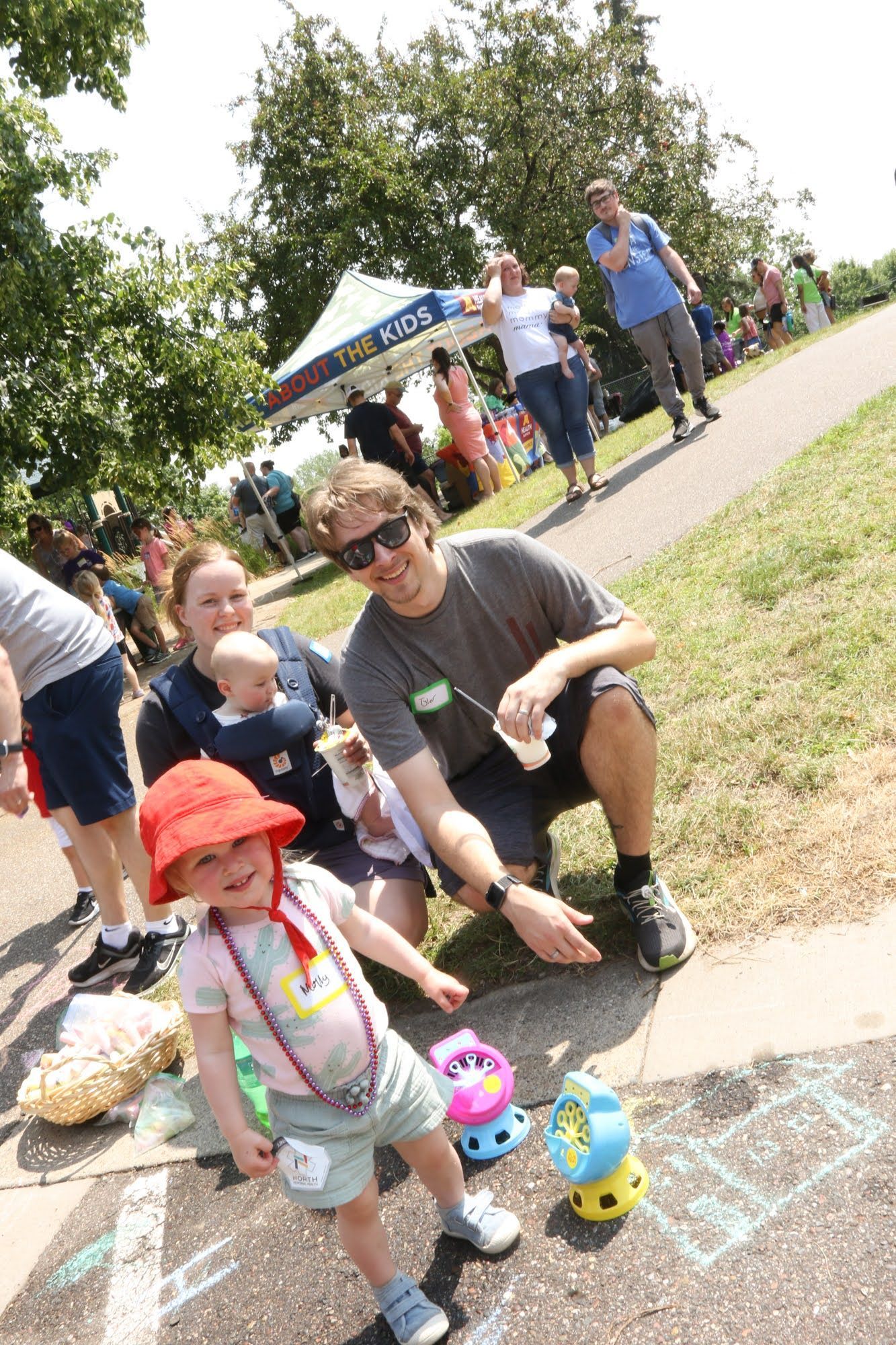 Family at an outdoor event with a young child in a red hat pointing at chalk drawings on the ground.