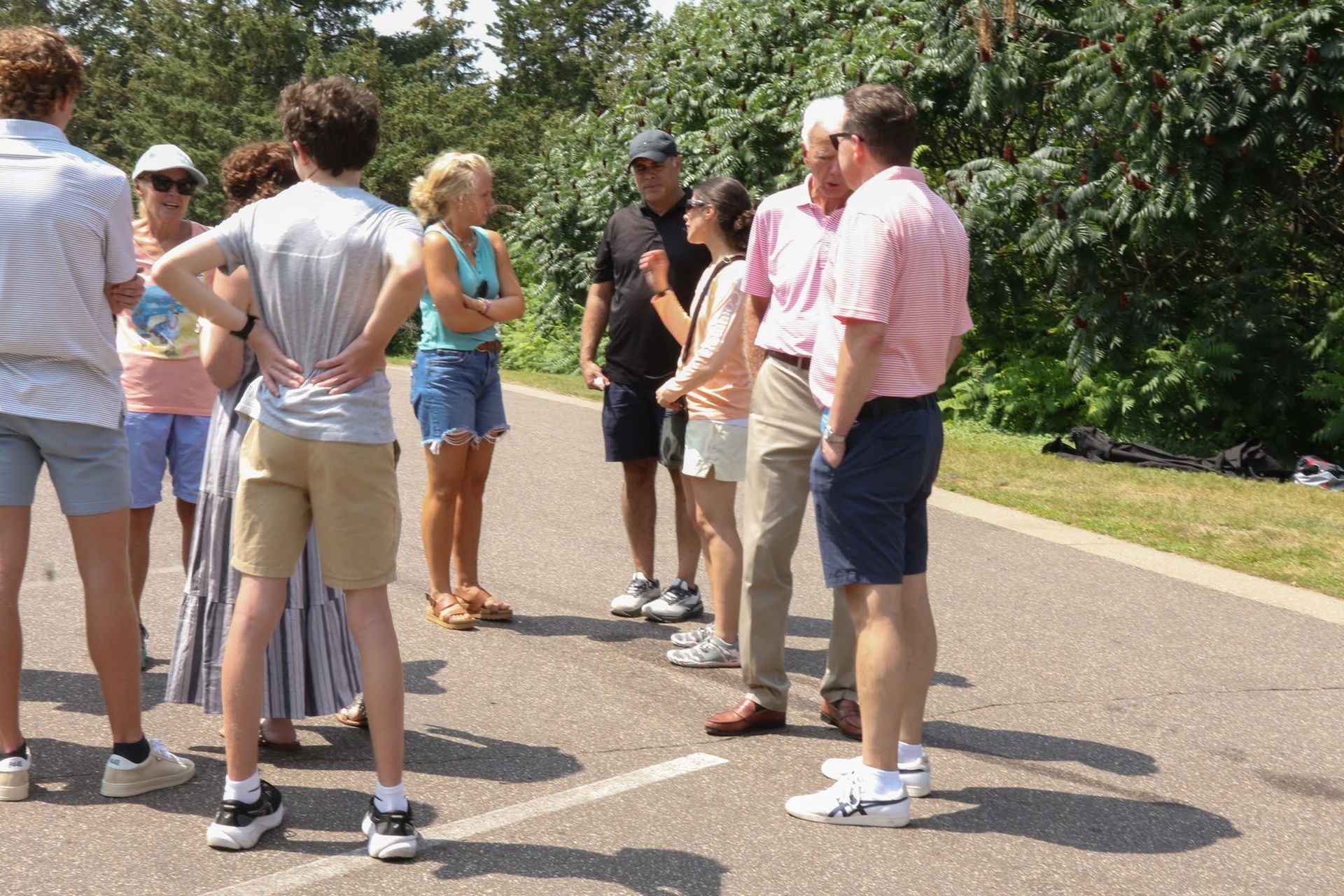 A group of people standing and talking on a paved path outdoors, under the sun.