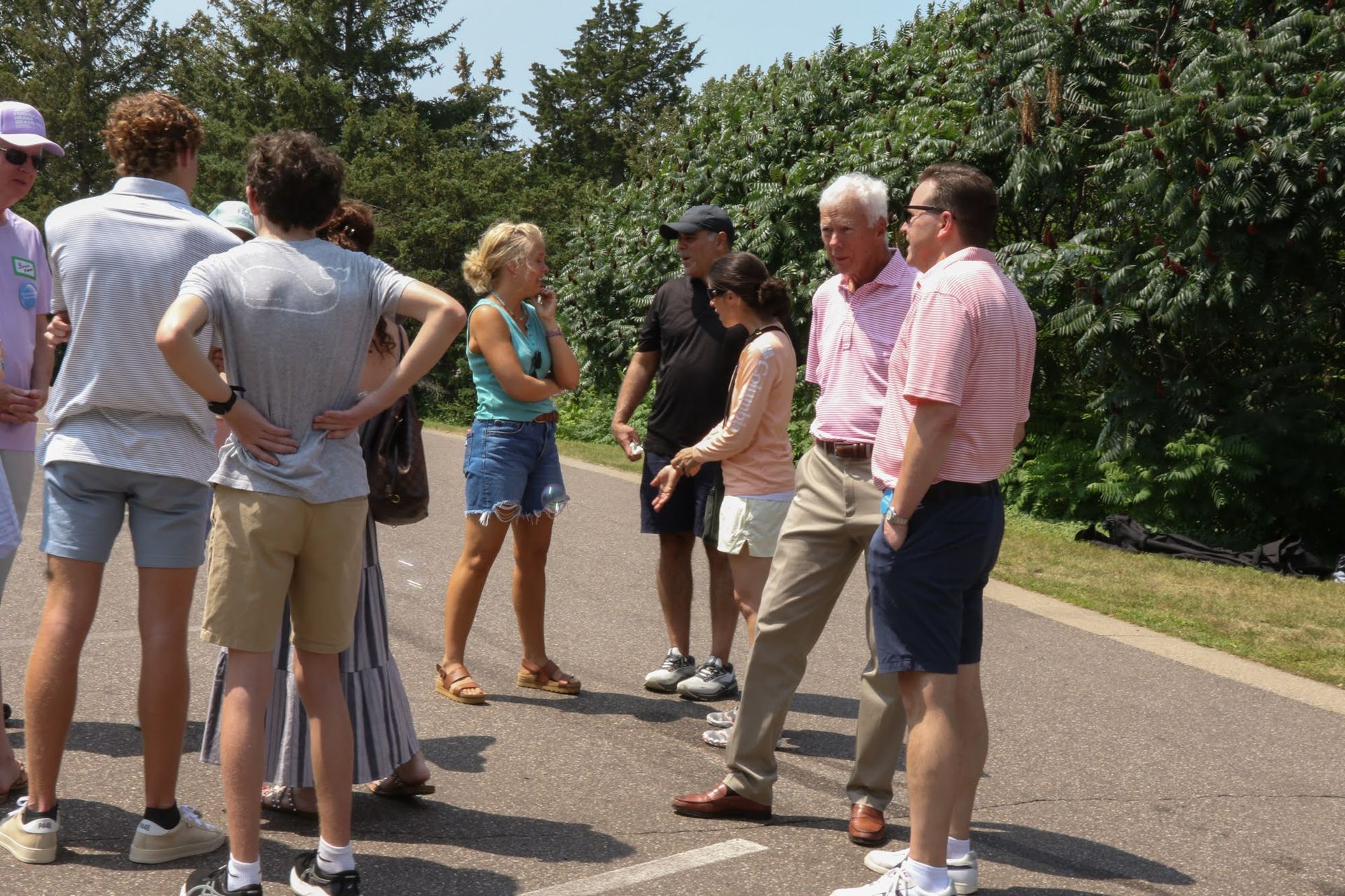 Group of people in casual attire outdoors, talking and standing on pavement in front of trees.