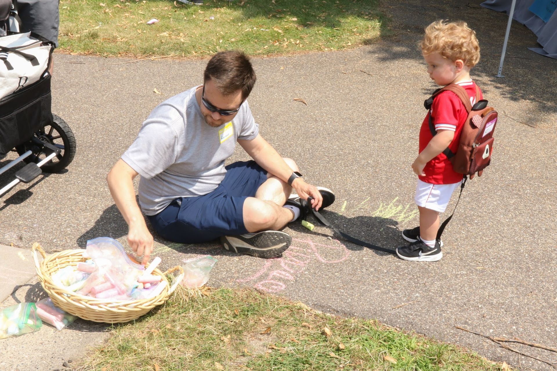 Man and child drawing with chalk on pavement. Basket of chalk nearby. Sunny day.