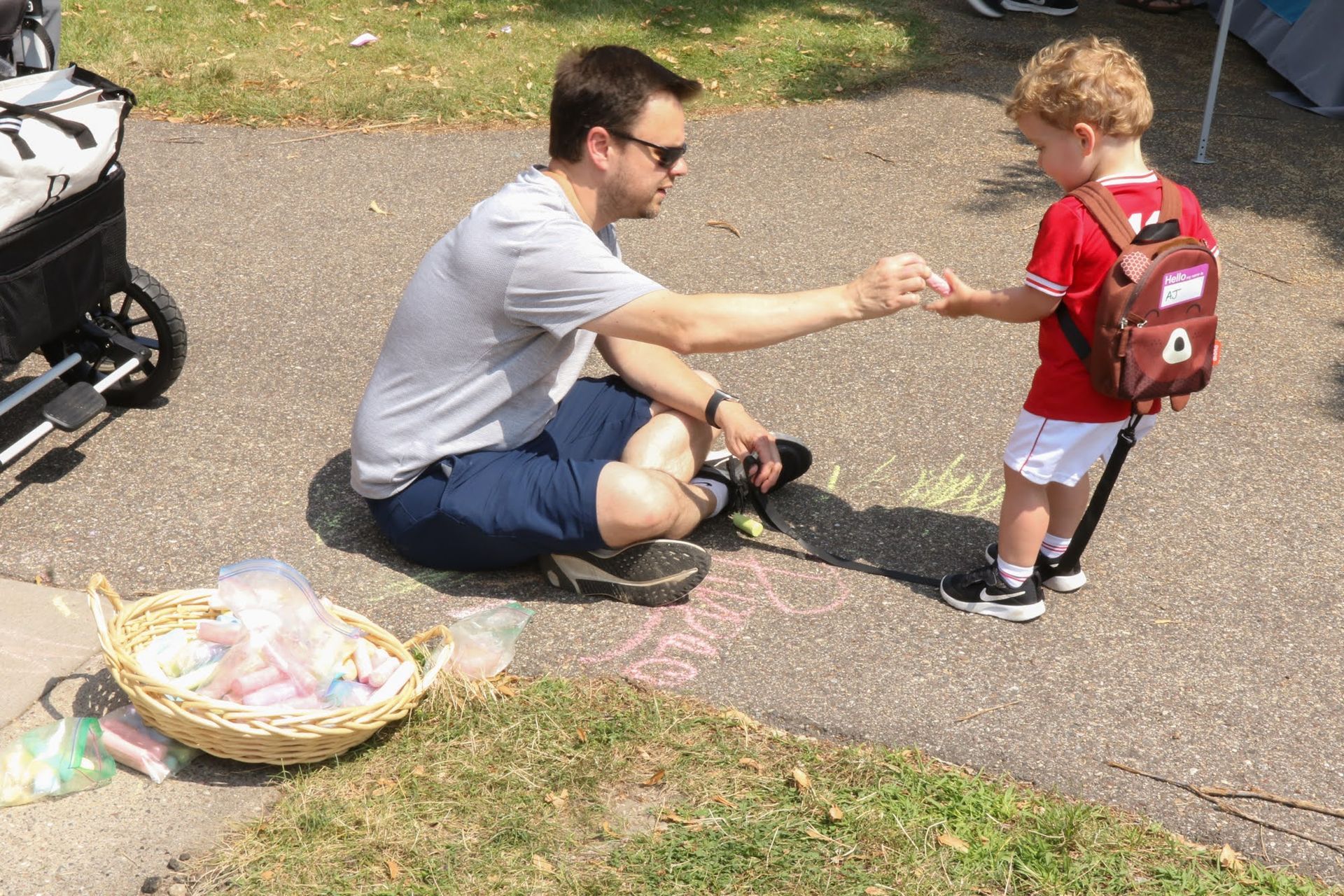 Man sitting, giving something to child wearing a backpack. Chalk drawings and basket of candy next to them. Outdoors, sunny.