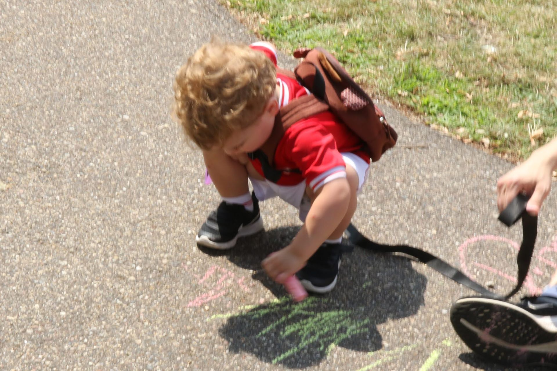 Child in red shirt and backpack crouching, drawing with chalk on a sidewalk.