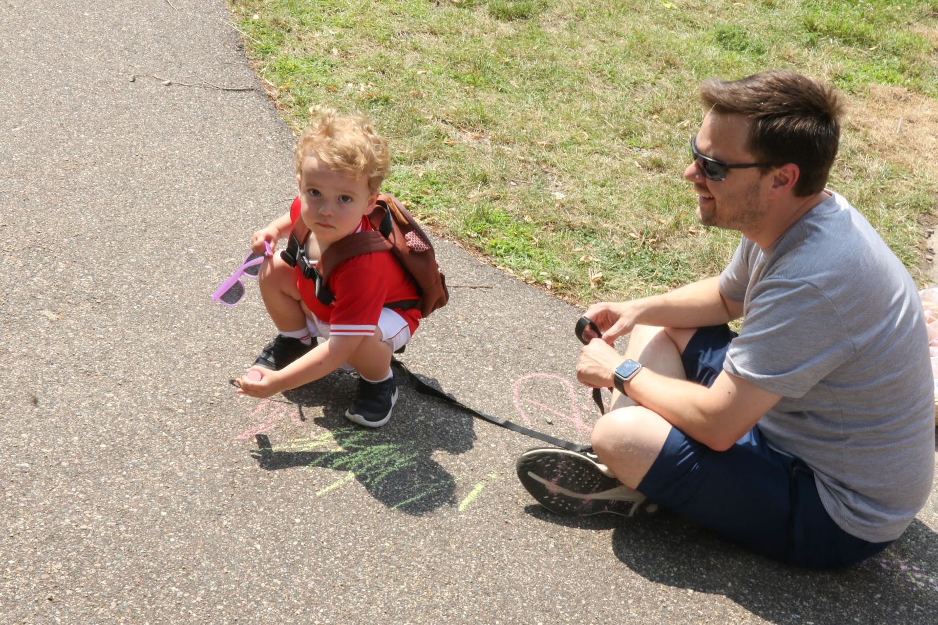 Child in red shirt and backpack squats drawing with chalk as man in gray shirt sits nearby.