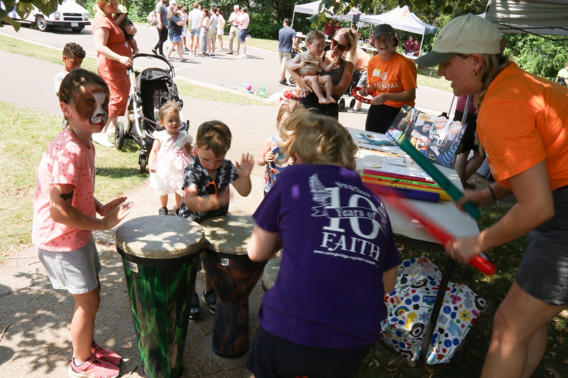 Children drumming at a park event, supervised by adults. People in the background. Sunny day.