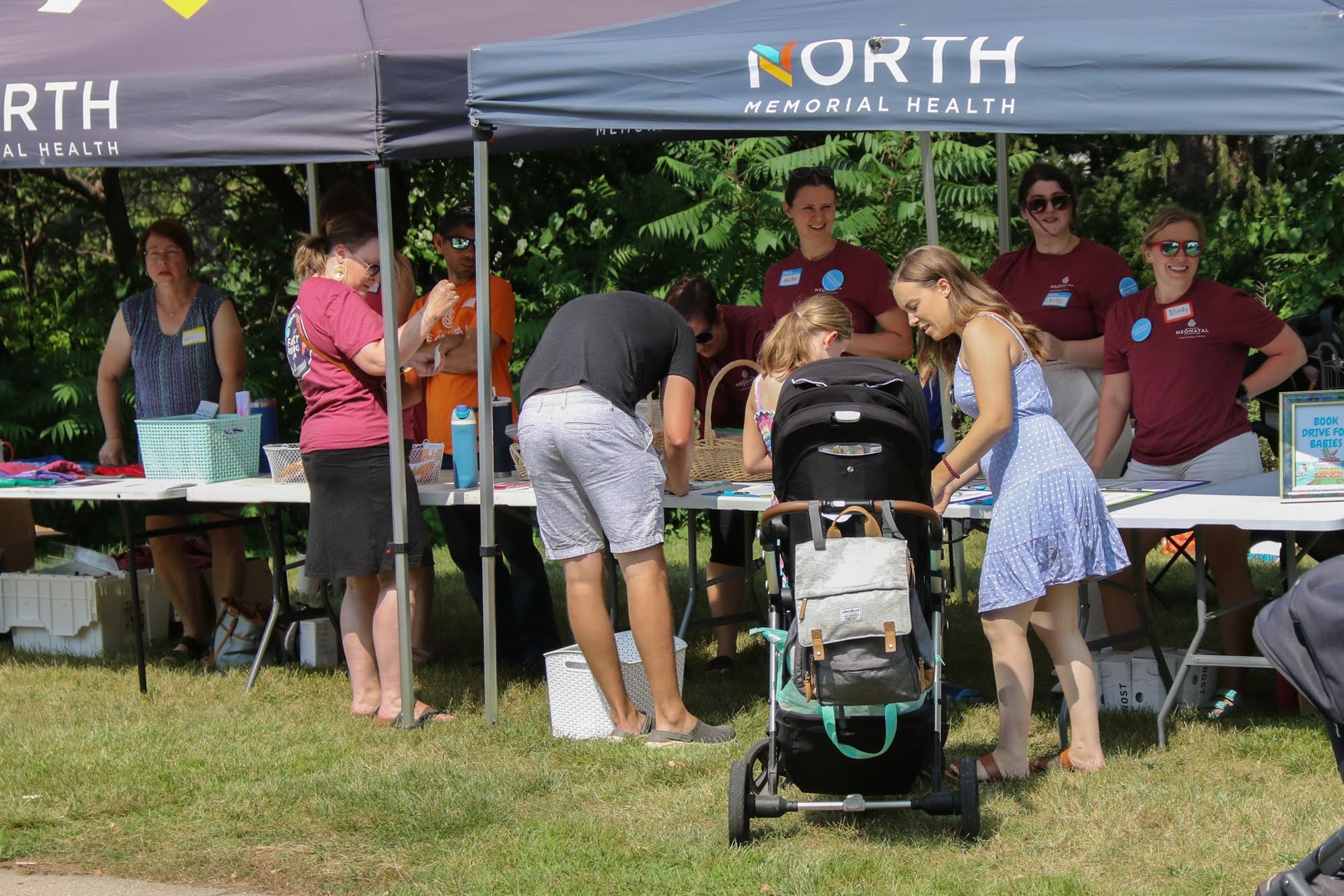 People at a North Memorial Health tent outdoors, signing up at a table; a stroller is in front.