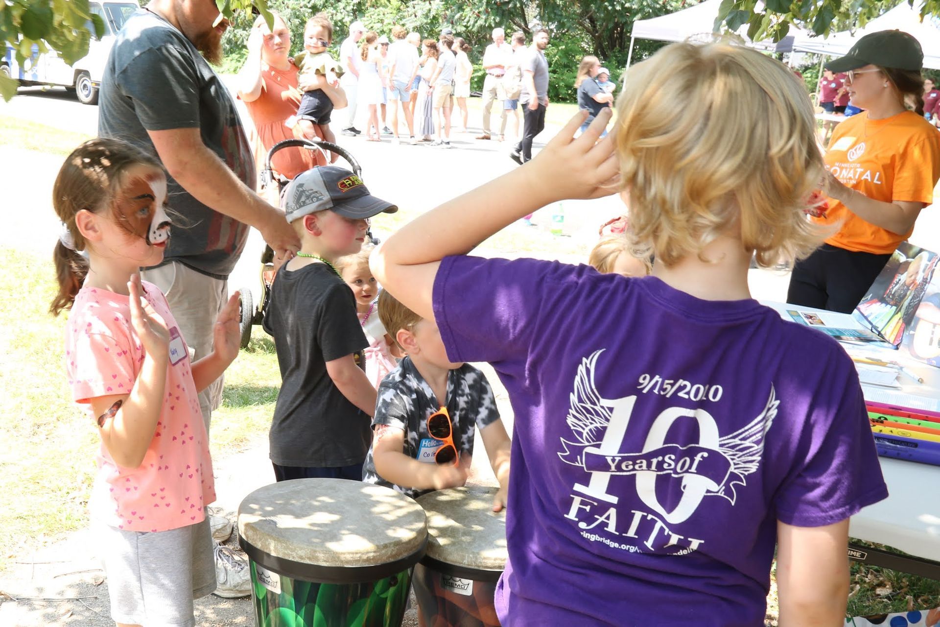 Children playing drums at an outdoor event; others watching. One child has face paint. Sunny day.