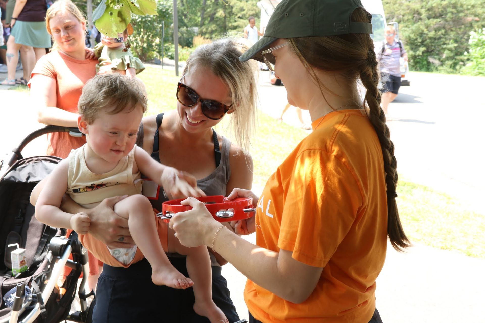 Woman in orange shirt offers a toy to a toddler held by a smiling woman; other people are nearby in a park setting.