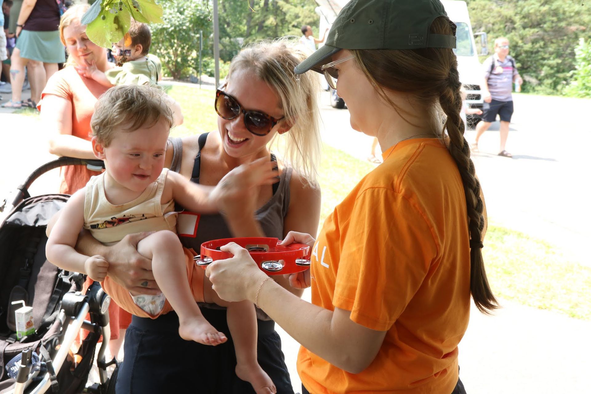 Woman holding a child and laughing as a person in an orange shirt offers a tambourine outdoors.