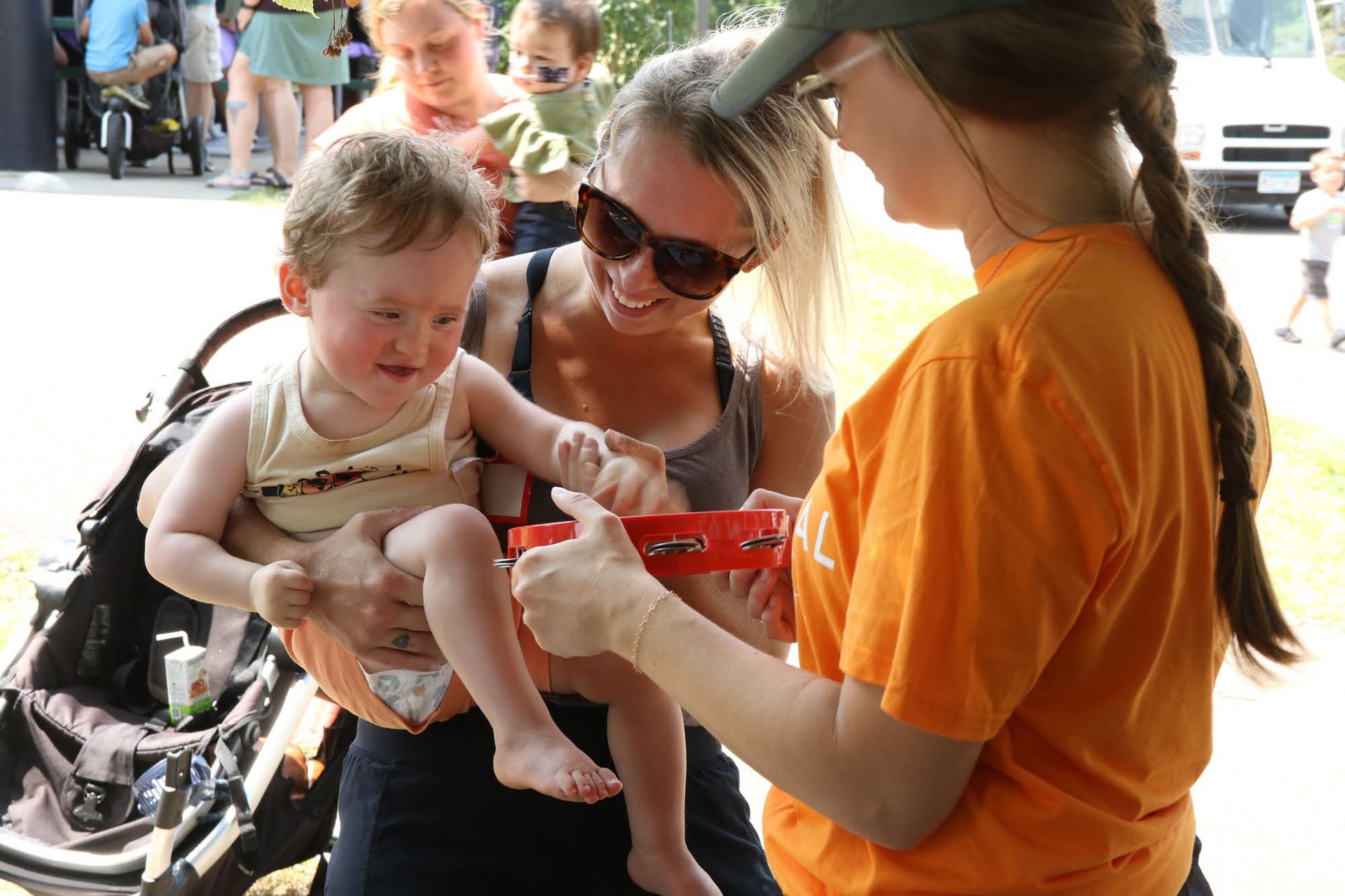 Woman in sunglasses holds a baby as another woman in an orange shirt shows the baby a red tambourine outdoors.