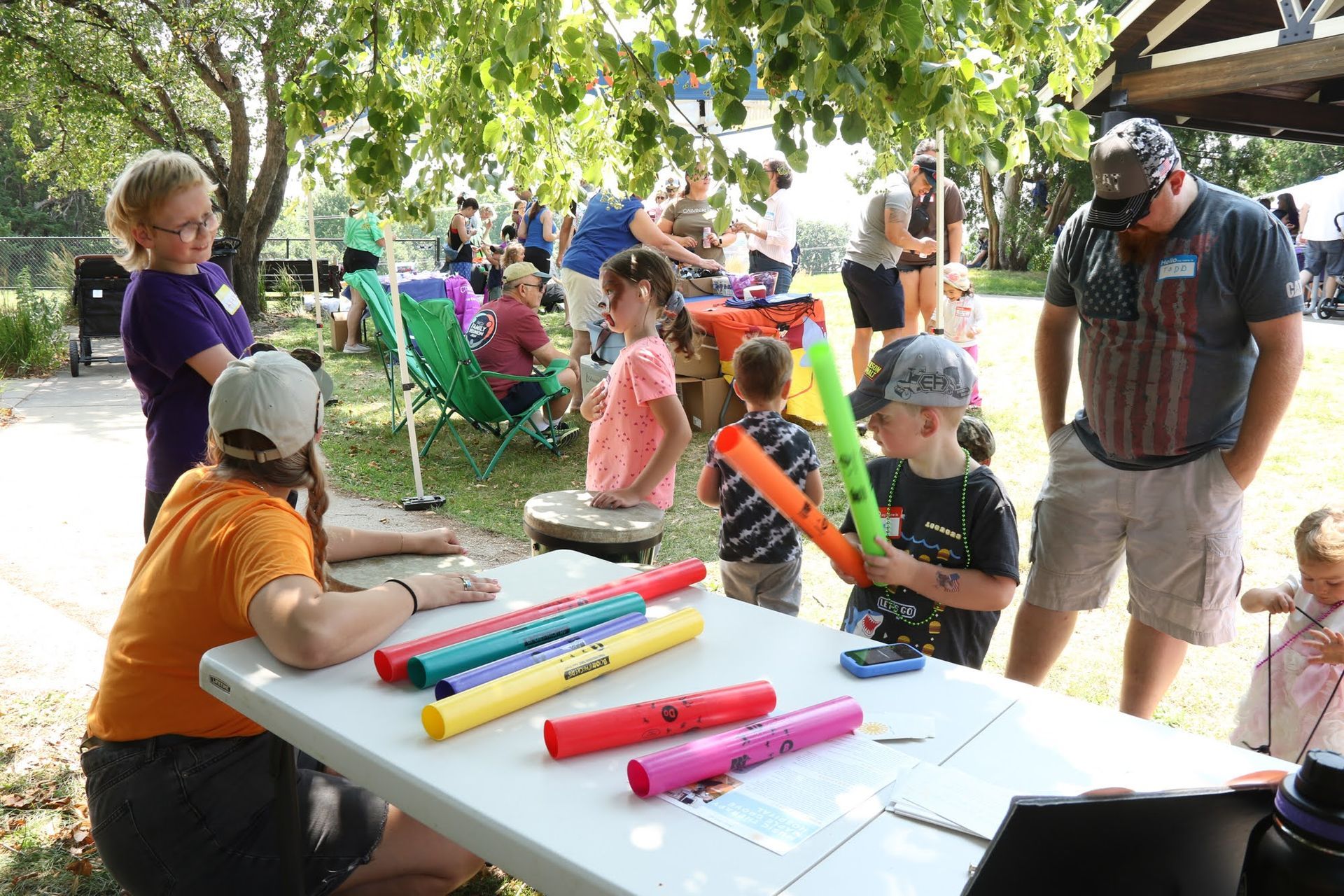 People at an outdoor event, including children playing with colorful musical tubes, near tables.