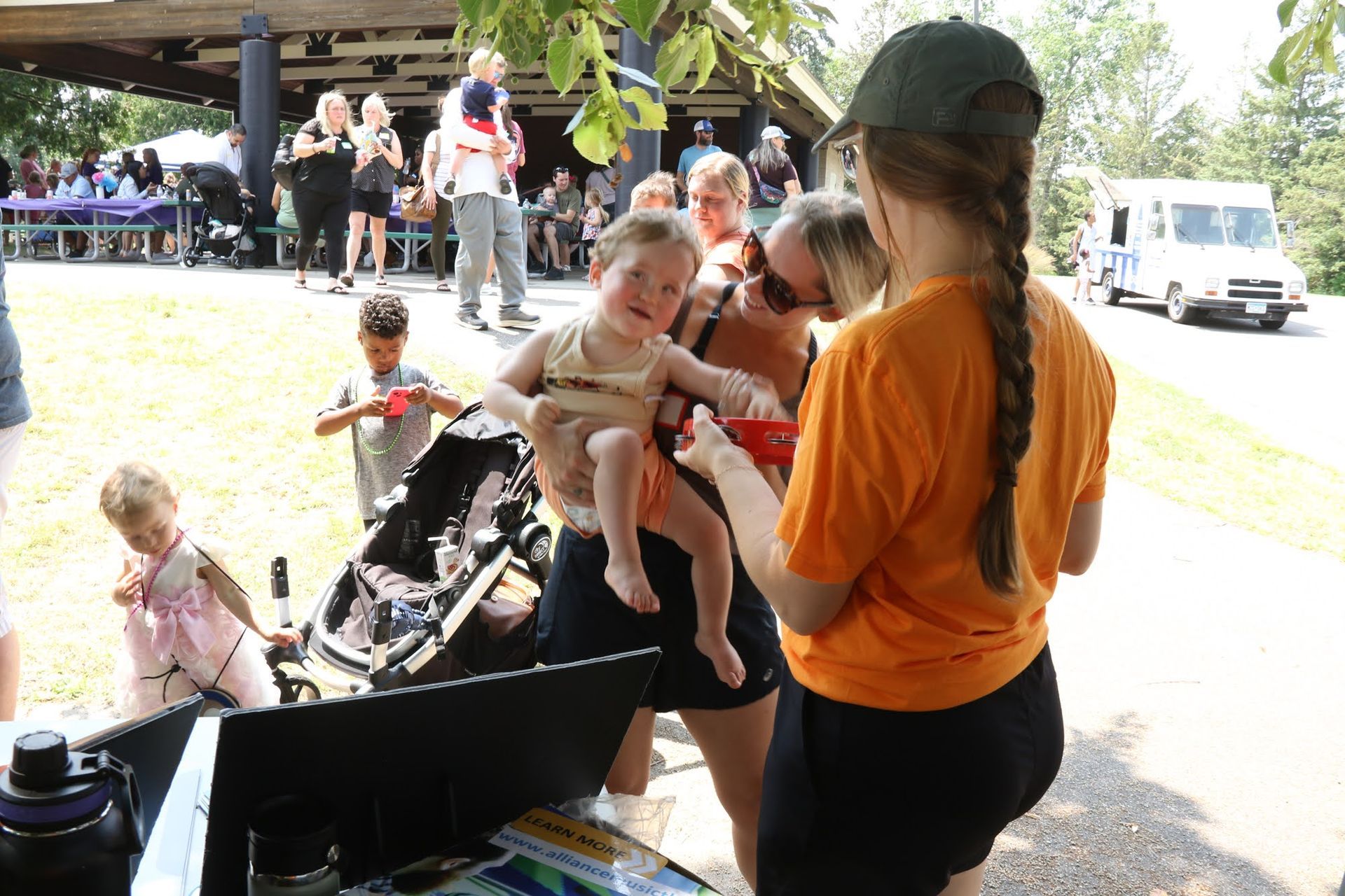 People at an outdoor event with a food truck, a child being held and receiving something from a person in an orange shirt.