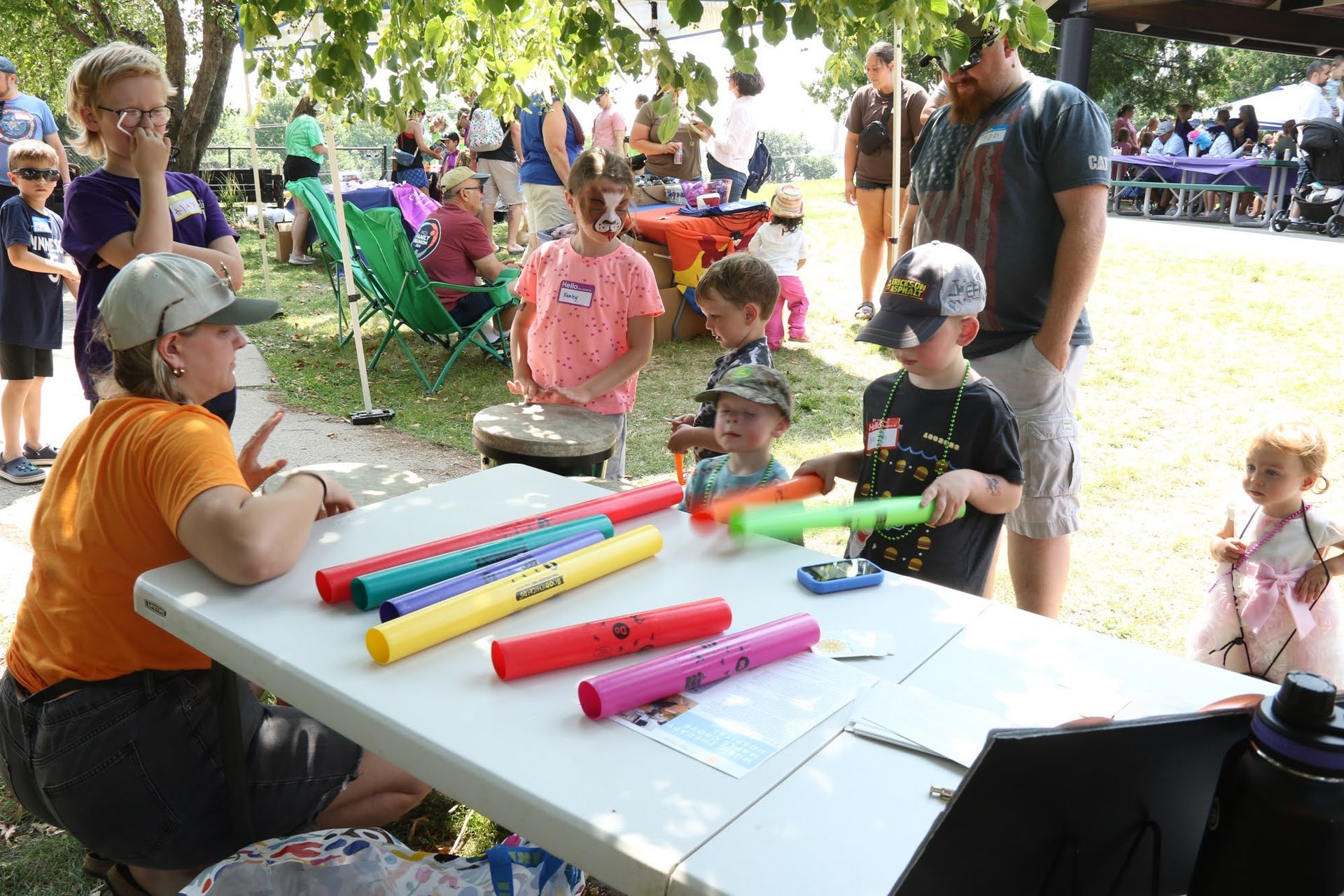 Children at a park table playing colorful musical instruments with an adult. Other people watch.