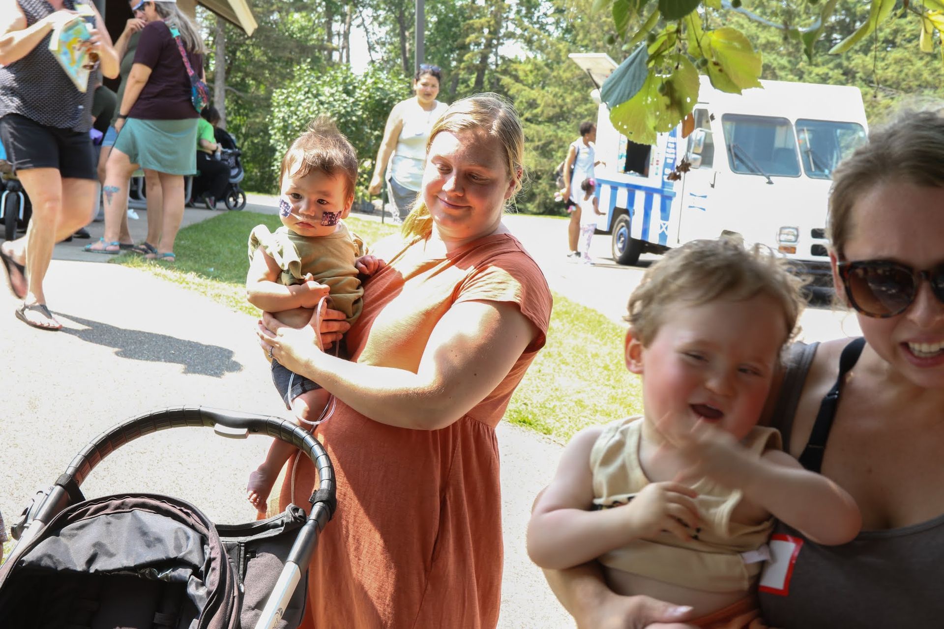 Woman holds a baby outdoors, another child nearby, food truck in the background, a stroller, sunny day.