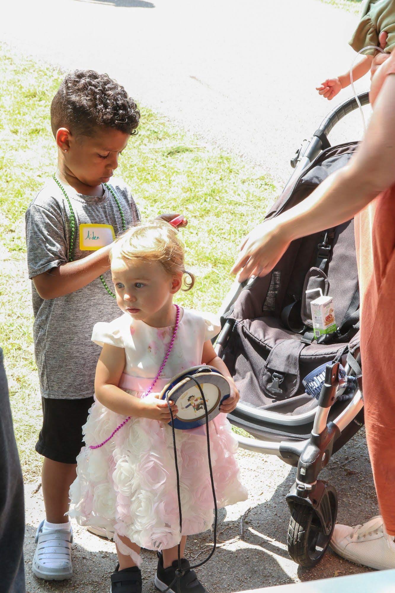 Boy with curly hair braids a girl's hair, holding a toy drum, near a stroller. Outdoors.