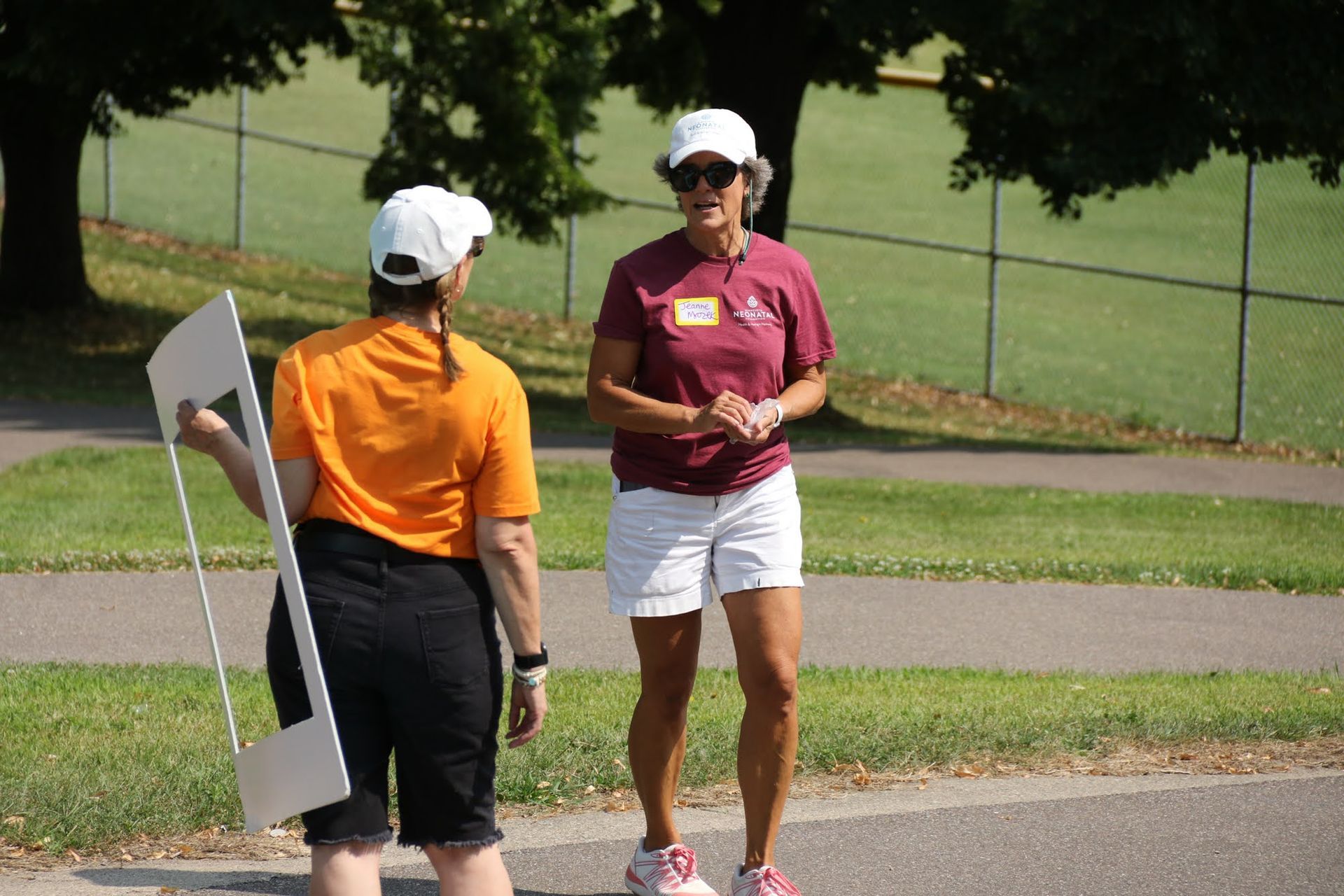 Two women in hats conversing outdoors near a fence. One holds a white sign, the other wears a maroon shirt.