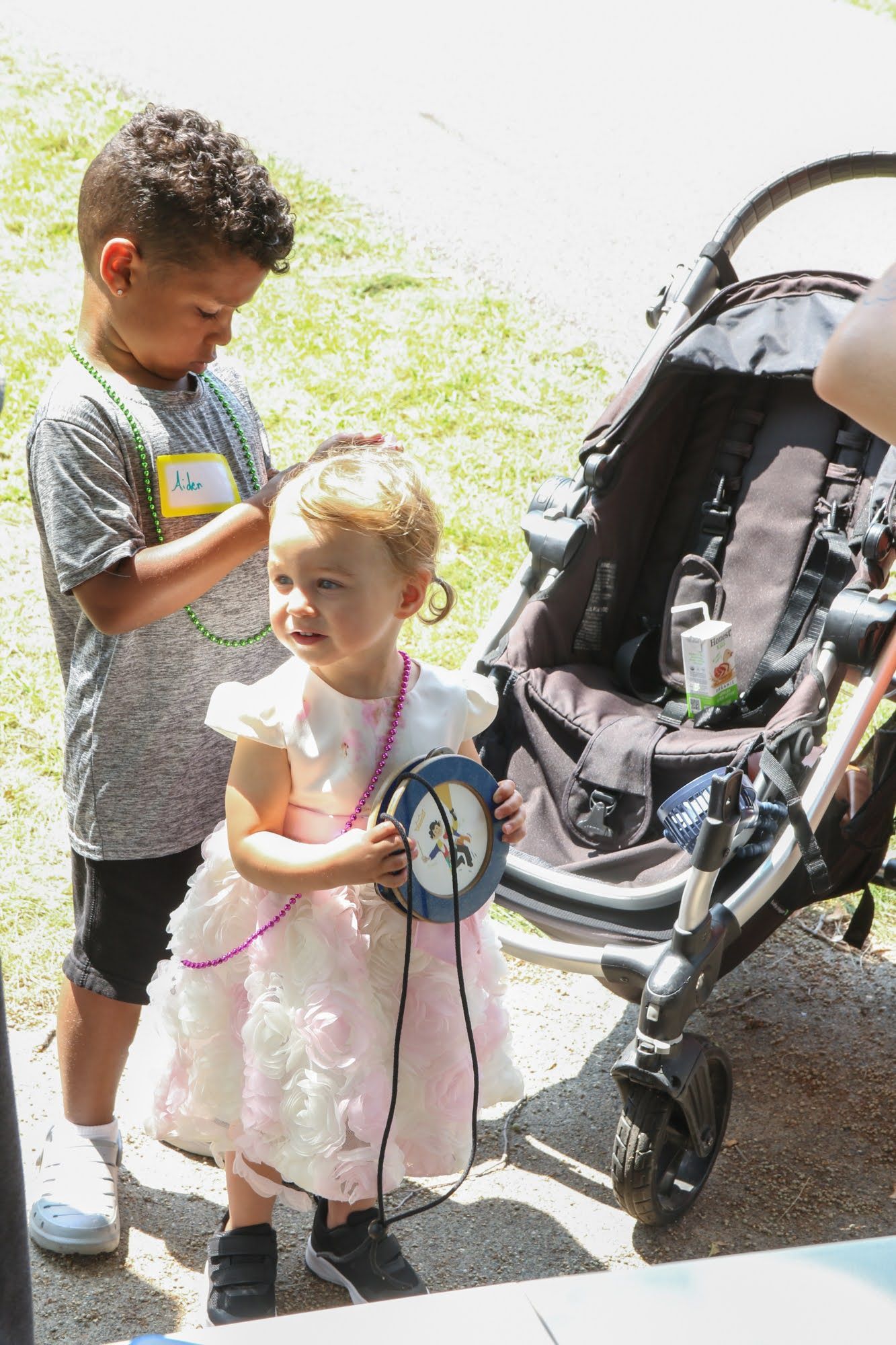 Two children near a stroller. One child is wearing a dress and holding a round object, the other is wearing a shirt and shorts.