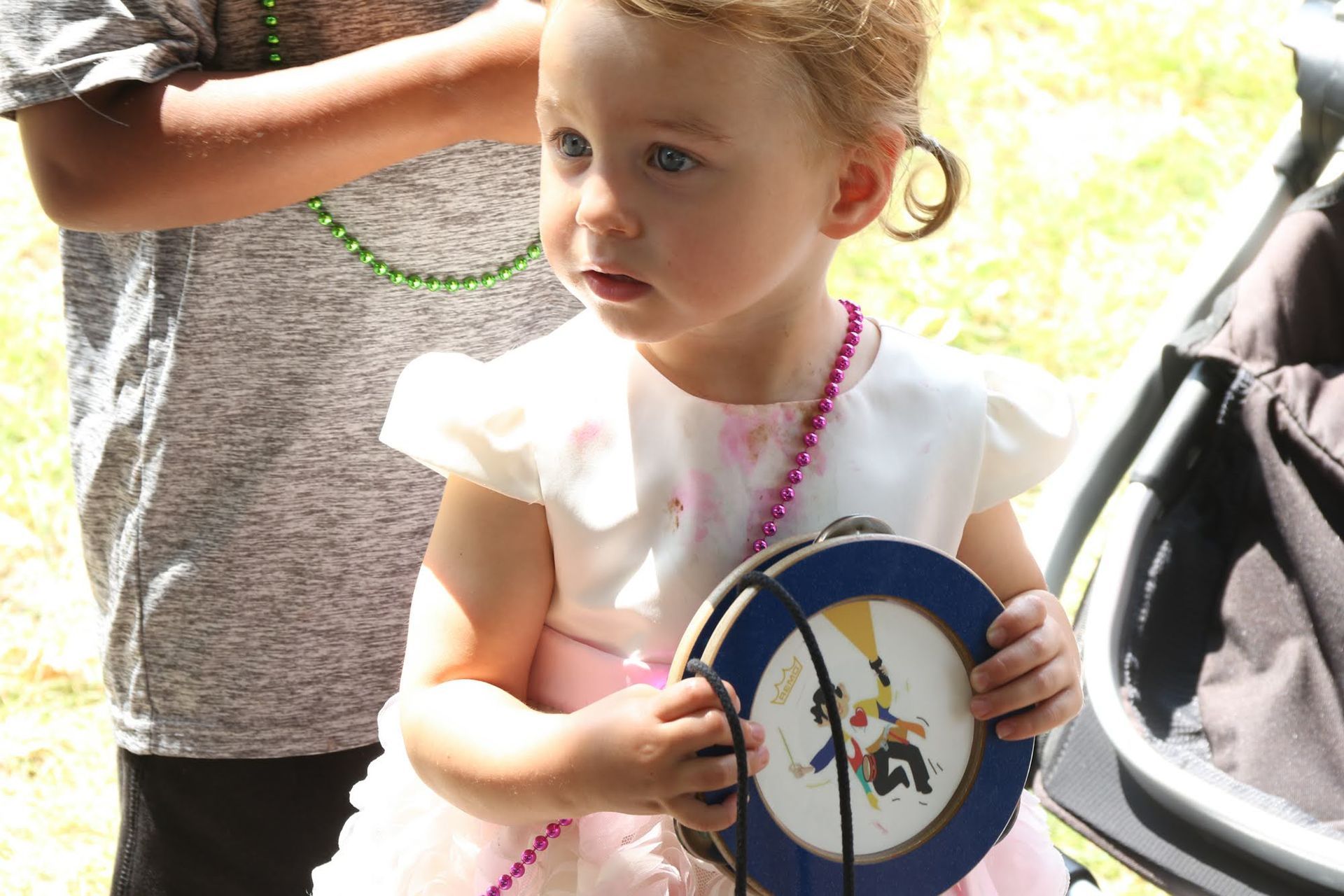 Young child with fair skin, wearing a pink dress, holding a tambourine and looking to the side.