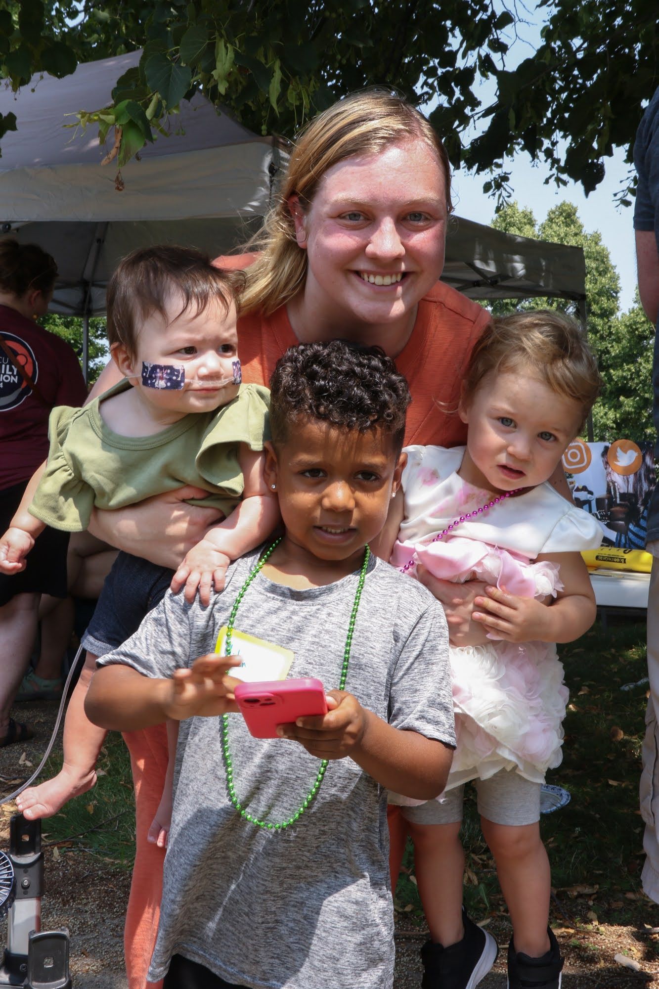 Woman holding two babies smiles with a young child who holds a small red object. Outside.