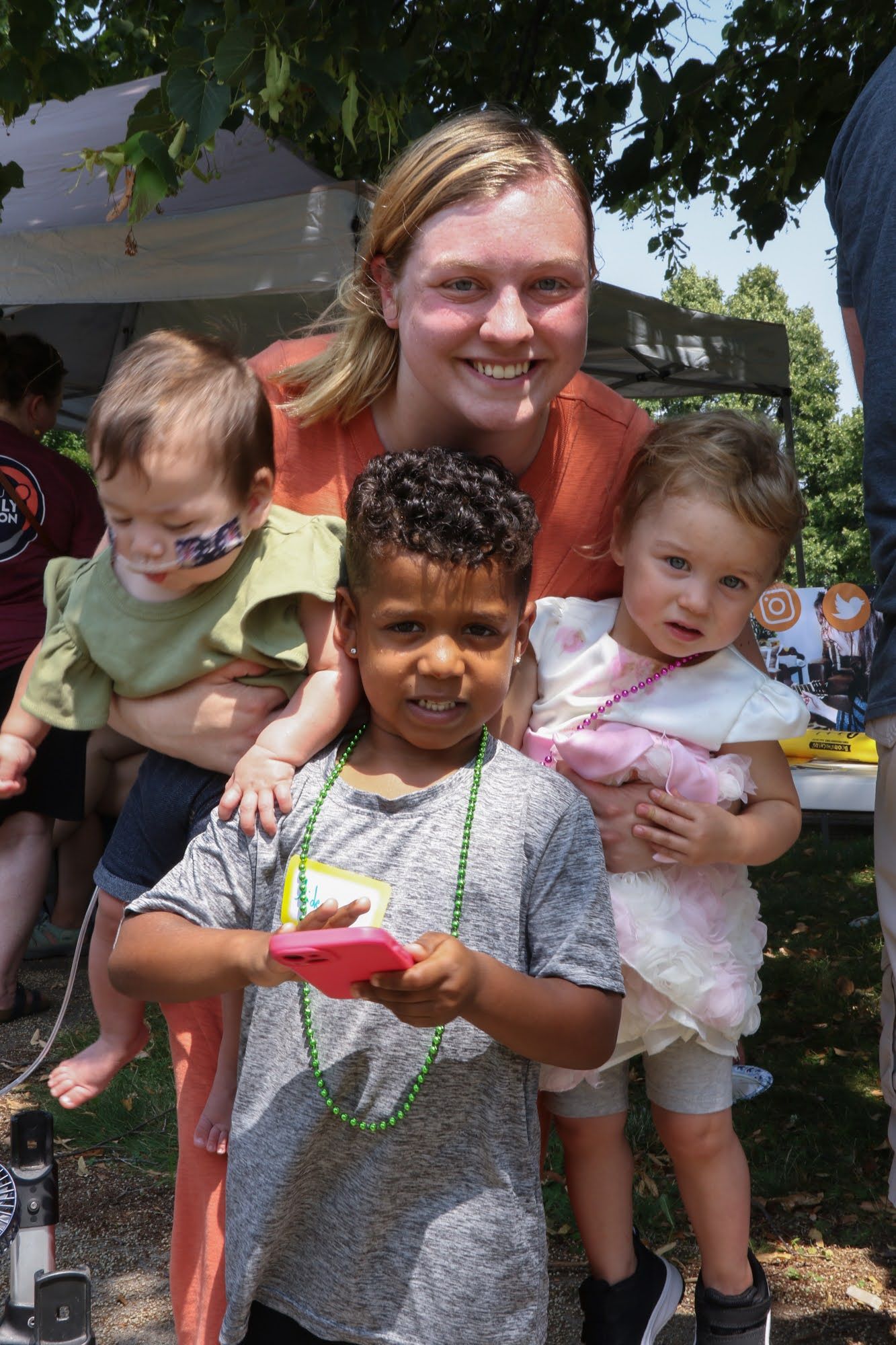 Woman holding two toddlers, smiling; another child in front holding a pink phone. Outdoors.