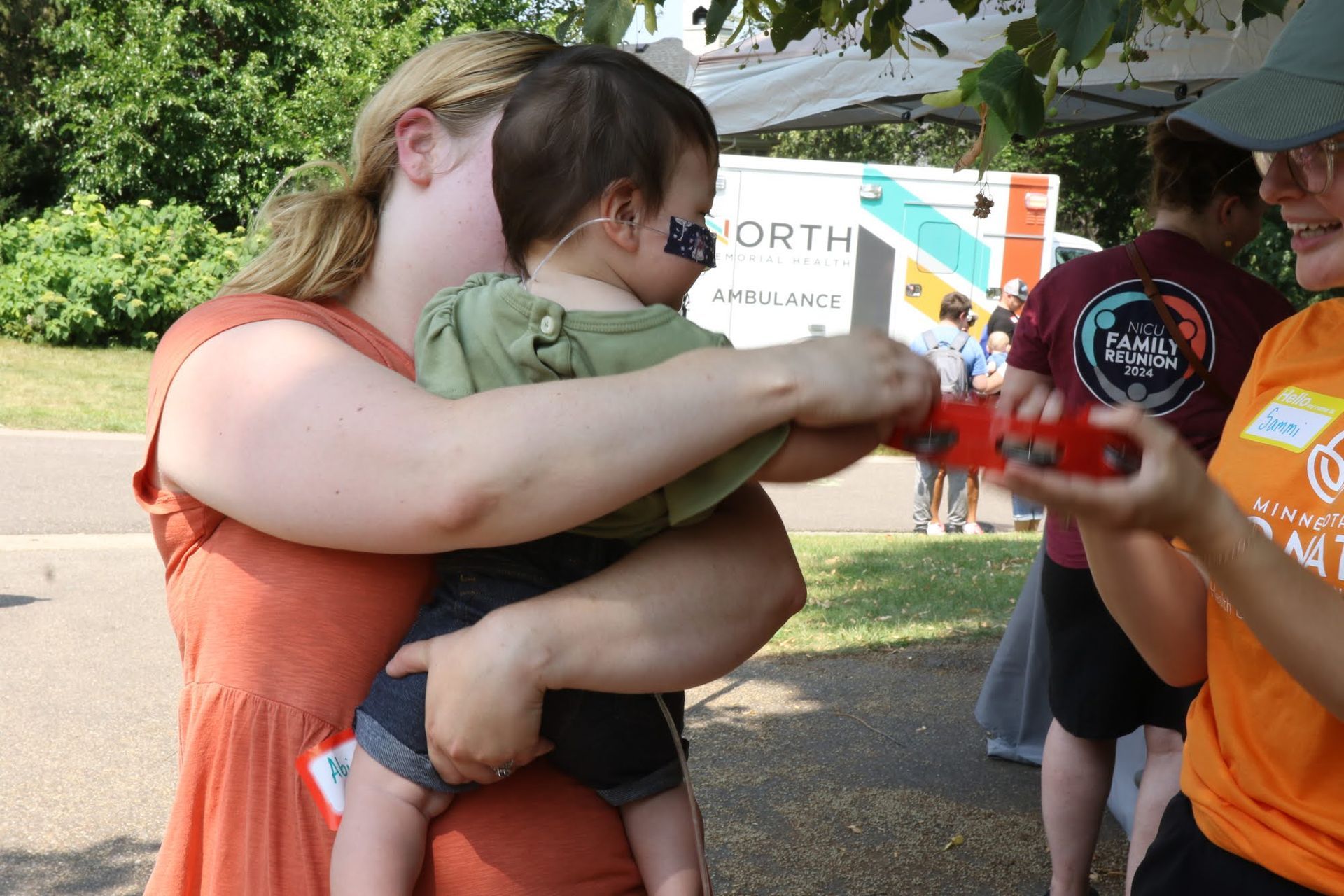 Woman holding a child; another person hands the child an object outdoors.