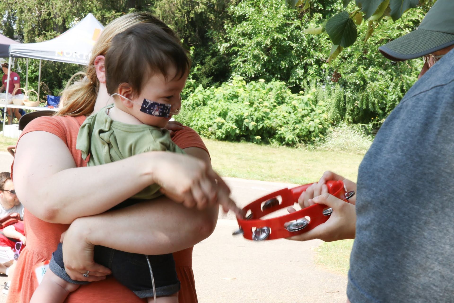 Woman holding a baby with a temporary face tattoo; another person offers a tambourine outdoors.