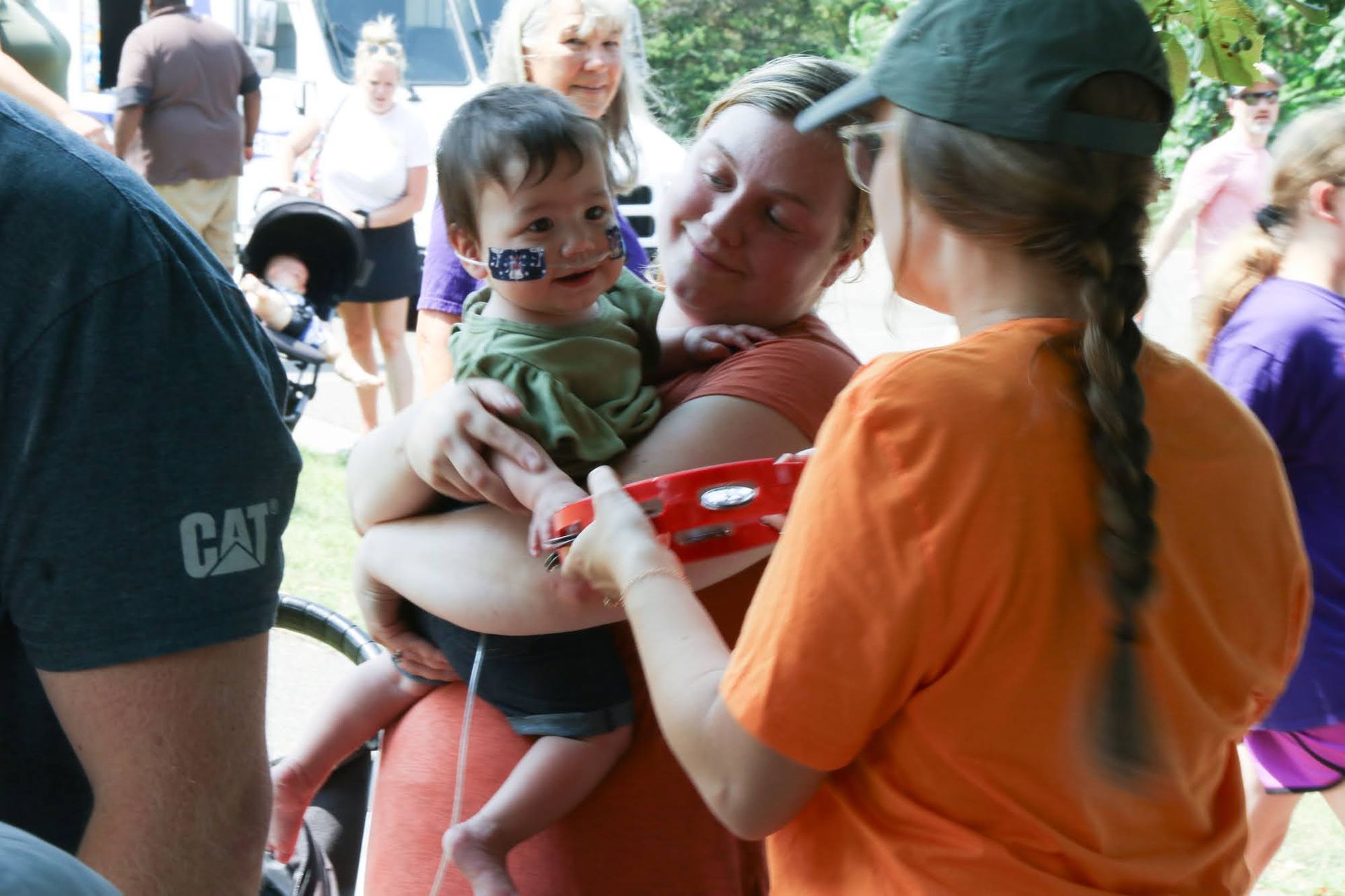 Woman holding baby with face paint, being given a toy by another person wearing an orange shirt and green cap, at an outdoor event.