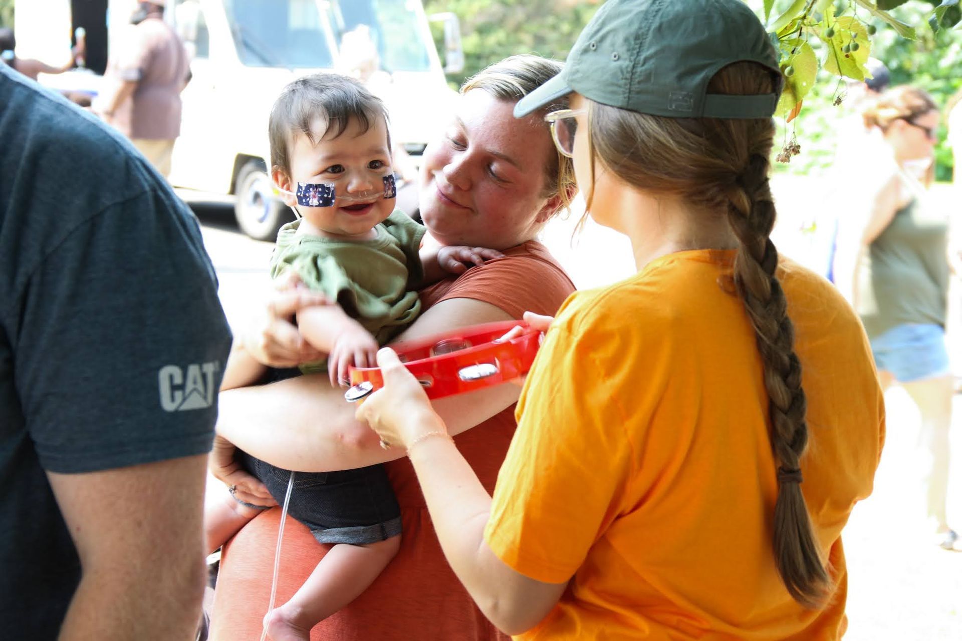 Woman holding a smiling baby with face paint, another woman offers a toy at an outdoor event.
