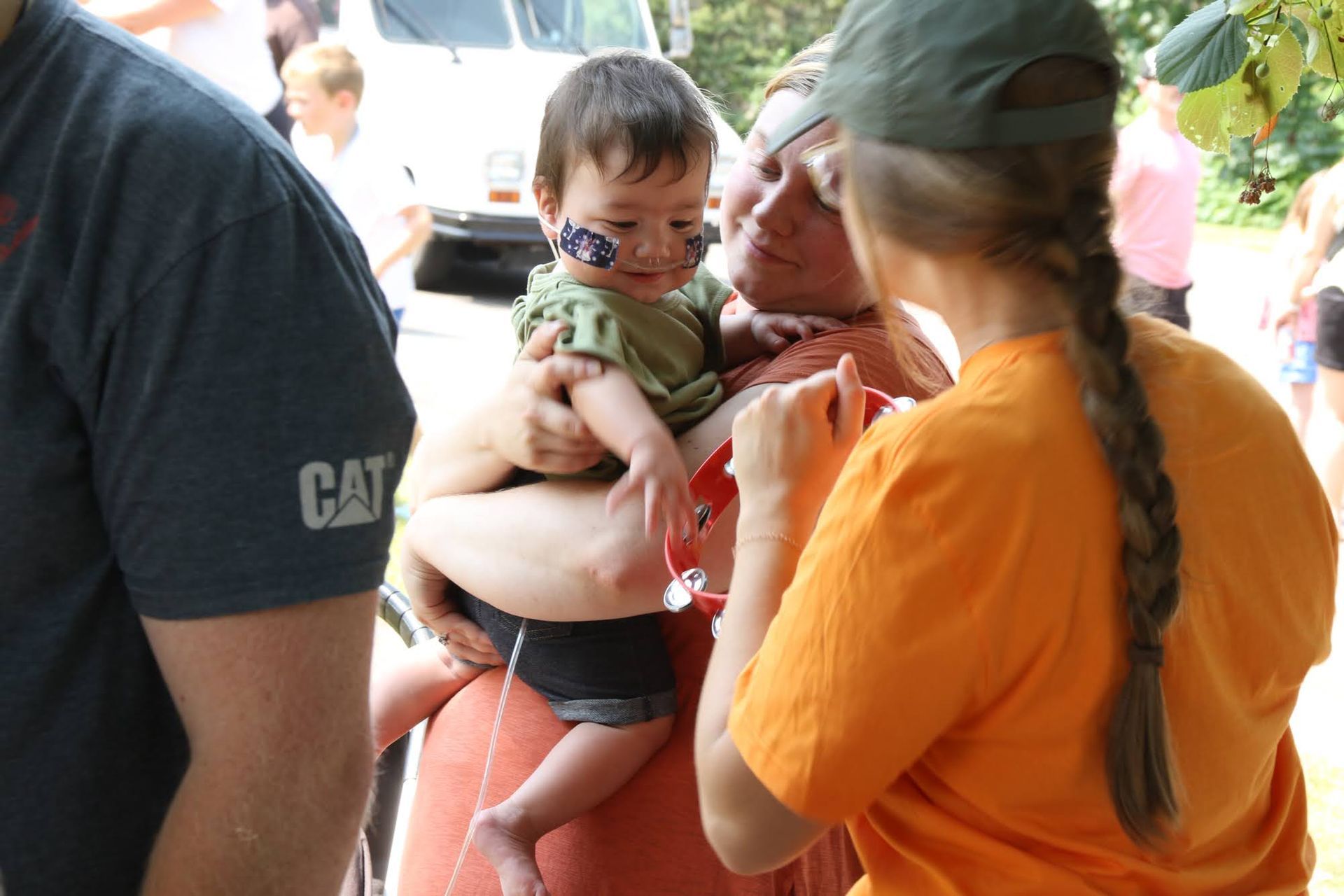 A person holding a child with face paint. A person is handing a cup to the child. Outdoors.