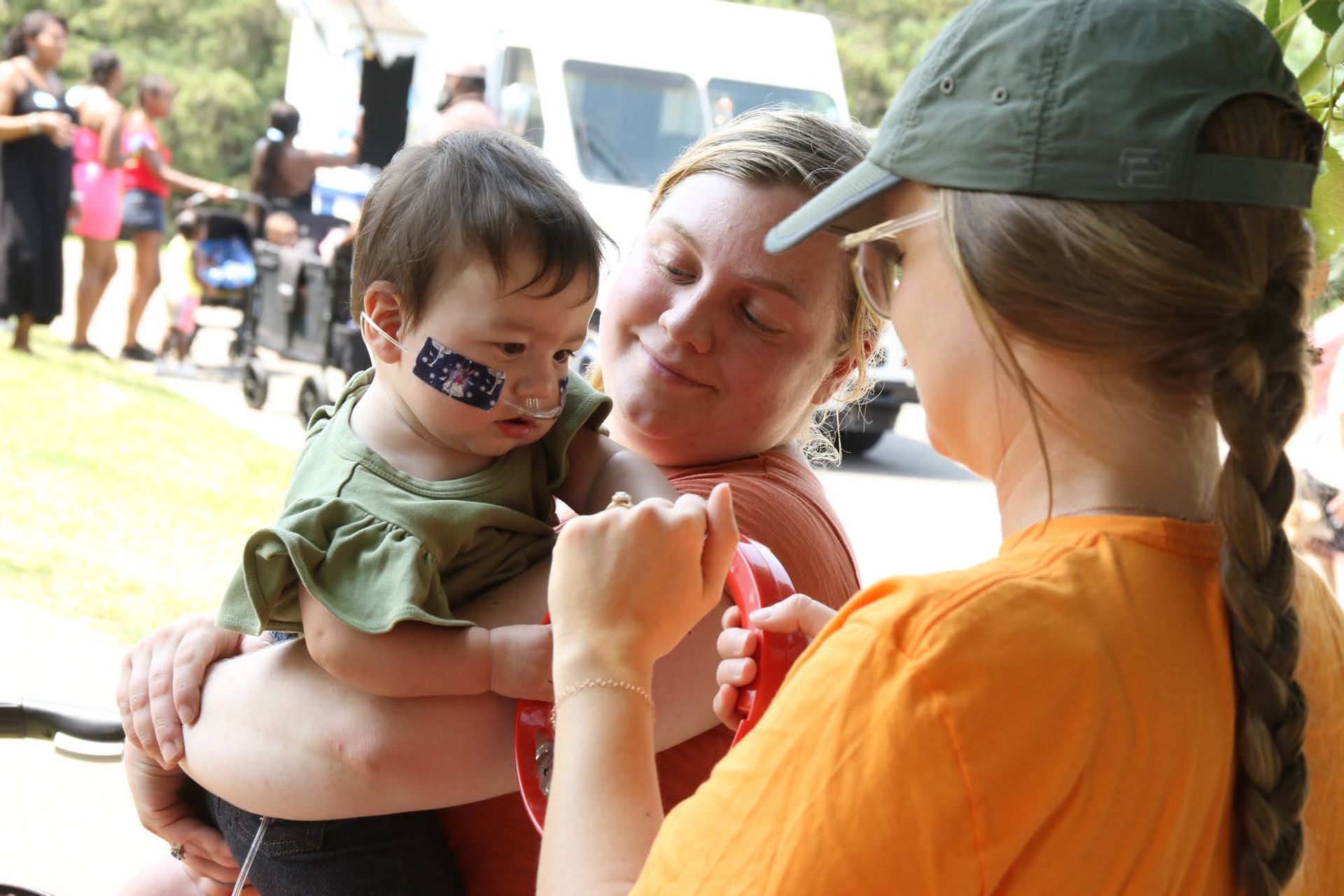 Woman holding child with face paint, another woman adjusts a red leash. Outdoor setting.