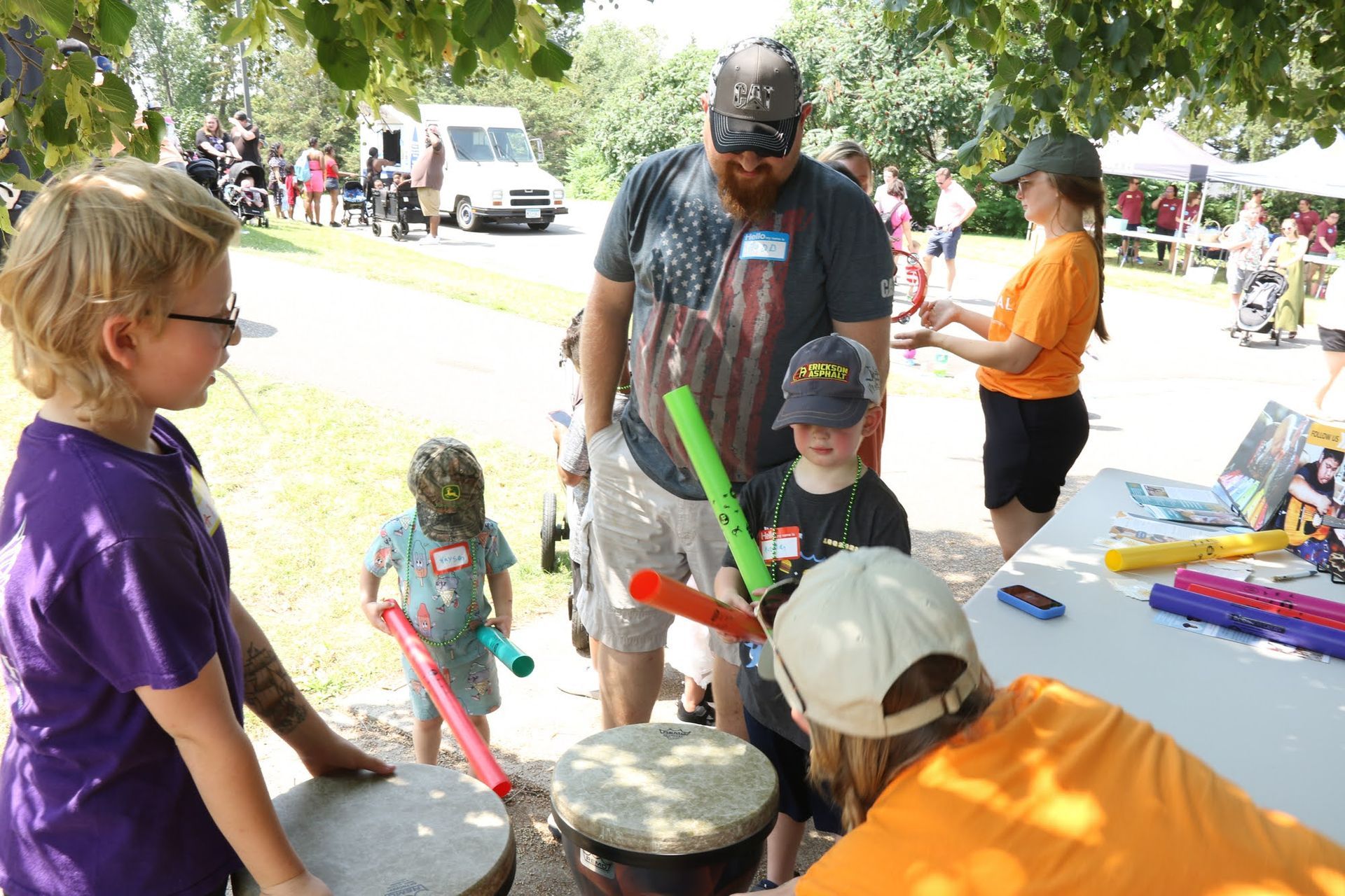 Children and adults at an outdoor event playing drums with sticks.