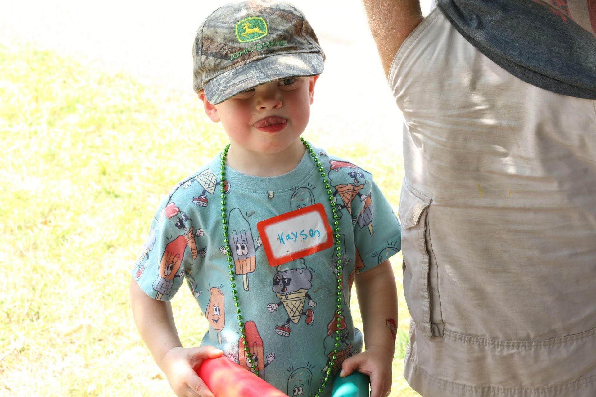 Boy wearing a camouflage hat and beads, holding red and green items, with a name tag that reads 